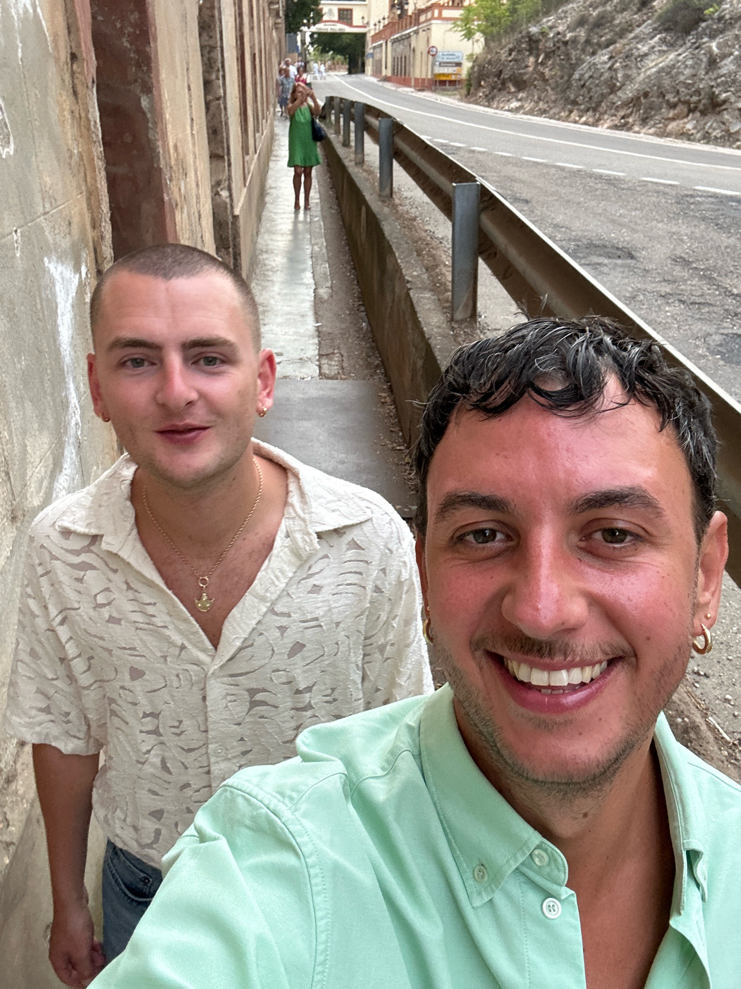 In Alhama de Aragón, Spain, two men are captured in a selfie taken from a low angle. The man in the foreground, on the right, has dark, curly hair and is wearing a light green button-down shirt. He is smiling broadly, revealing white teeth. His right earlobe features a gold hoop earring. To his left, the second man has shorter, light brown hair and is wearing a textured, light-colored button-down shirt with a gold chain necklace featuring a pendant. He also appears to be smiling. They are positioned on a narrow pedestrian walkway bordered by a concrete wall on the left and a metal guardrail overlooking a road on the right. In the background, a woman in a green dress is visible further down the walkway, and buildings and a hillside are present beyond the road. The scene suggests an outdoor setting, possibly a street or a path in a town.