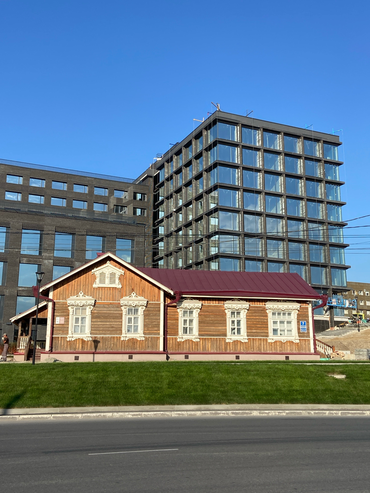 The image depicts a scene in Novosibirsk, Russia, featuring a contrast between traditional and modern architecture. In the foreground, a charming, well-maintained wooden house with ornate window surrounds and a dark red roof stands on a green lawn. The house has several windows, some with detailed decorative elements, and a small porch on the left side. To the right of the house, construction is underway, indicated by scaffolding and building materials.
Behind the wooden house, two large, contemporary buildings dominate the background. The building directly behind the house is a multi-story structure with a dark, textured facade and rows of windows. To its right, a more modern building with a glass facade, reflecting the blue sky and clouds, rises prominently. The glass building has a grid-like structure of dark frames surrounding numerous large windows.
The sky is a clear, vibrant blue, suggesting a sunny day. Power lines are visible stretching across the upper right portion of the image. A street with asphalt pavement and a concrete curb is in the lower part of the frame. A single person, dressed in casual attire, is visible on the left side of the wooden house. Another individual is partially visible on the right side near the construction area. The overall impression is a juxtaposition of historical charm and urban development in Novosibirsk.