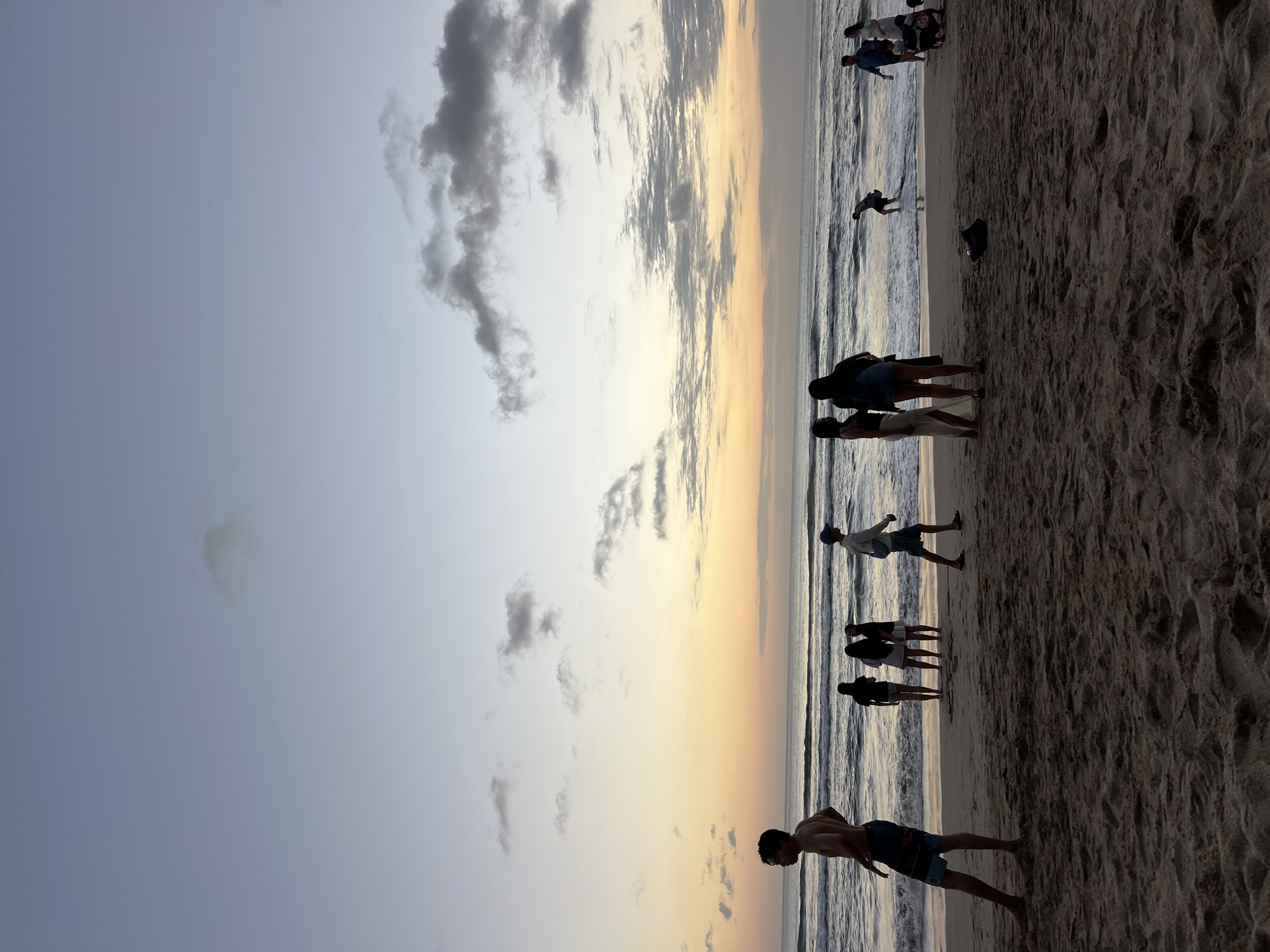 This is a photograph of a beach scene in Kuta, Indonesia, at sunset. The sky is a gradient of blue and orange, with scattered clouds. The ocean is calm, with gentle waves rolling onto the shore. Several people are visible on the beach, some walking, some standing, and others playing in the water. The sand is dark and textured, with footprints and ripples indicating recent activity. In the foreground, a man is standing facing left, with his back to the camera, wearing swim trunks. Further back, a group of people are gathered near the water's edge, some appearing to be talking or observing the sunset. The overall impression is one of a peaceful evening on a popular tourist beach.
