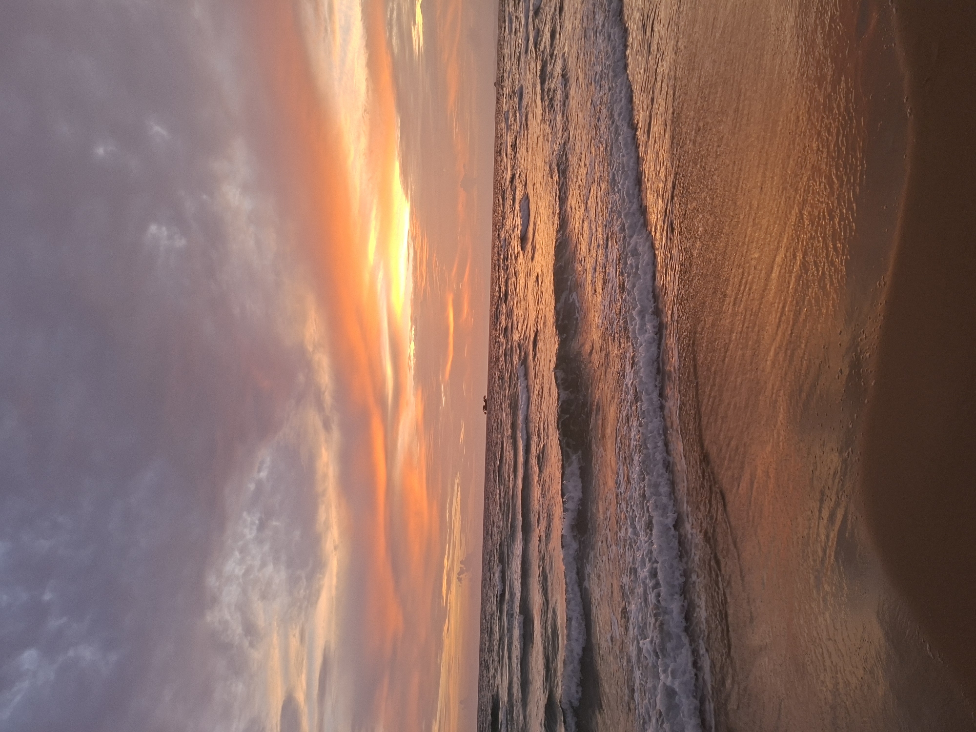 The image captures a sunset over the ocean in Phu Quoc, Vietnam. The sky is painted with vibrant hues of orange, pink, and purple, with clouds stretching across the horizon. Gentle waves lap at the sandy shore, reflecting the colors of the sunset. A distant boat can be seen on the water. The scene is tranquil and serene, evoking a sense of peace and beauty.