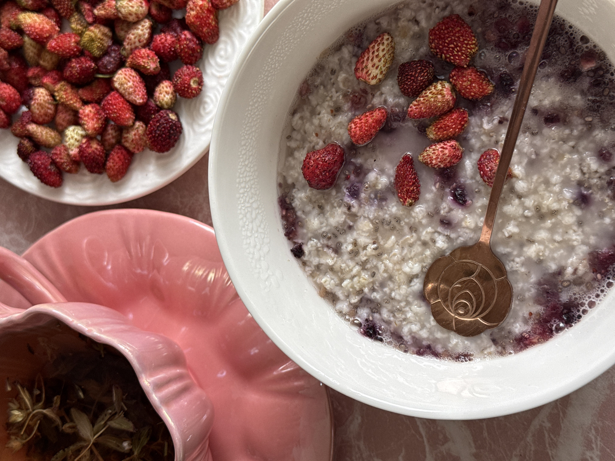 A close-up, high-angle shot shows a bowl of oatmeal topped with wild strawberries and a rose-shaped spoon. A pink teacup and saucer filled with tea, along with a plate of wild strawberries, are visible in the background. The scene is set on a pink marble surface.