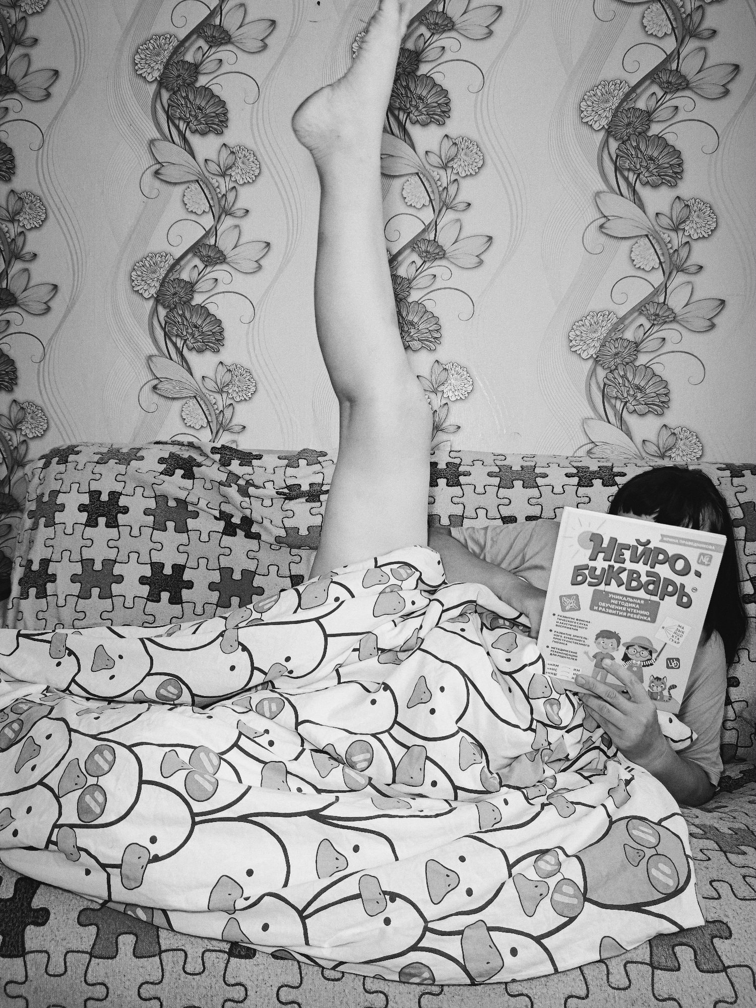 A black-and-white photograph depicts a person lying in bed reading a book. The person is mostly covered by a duvet patterned with cartoonish ducks, with one leg extended vertically into the air. The book, titled "НЕЙРО букварь" (Neuro Primer), is held in their hands. Behind the person, a pillow with a jigsaw puzzle pattern rests against a wall adorned with a floral wallpaper design. The photo was presumably taken in Фоки, Russia.