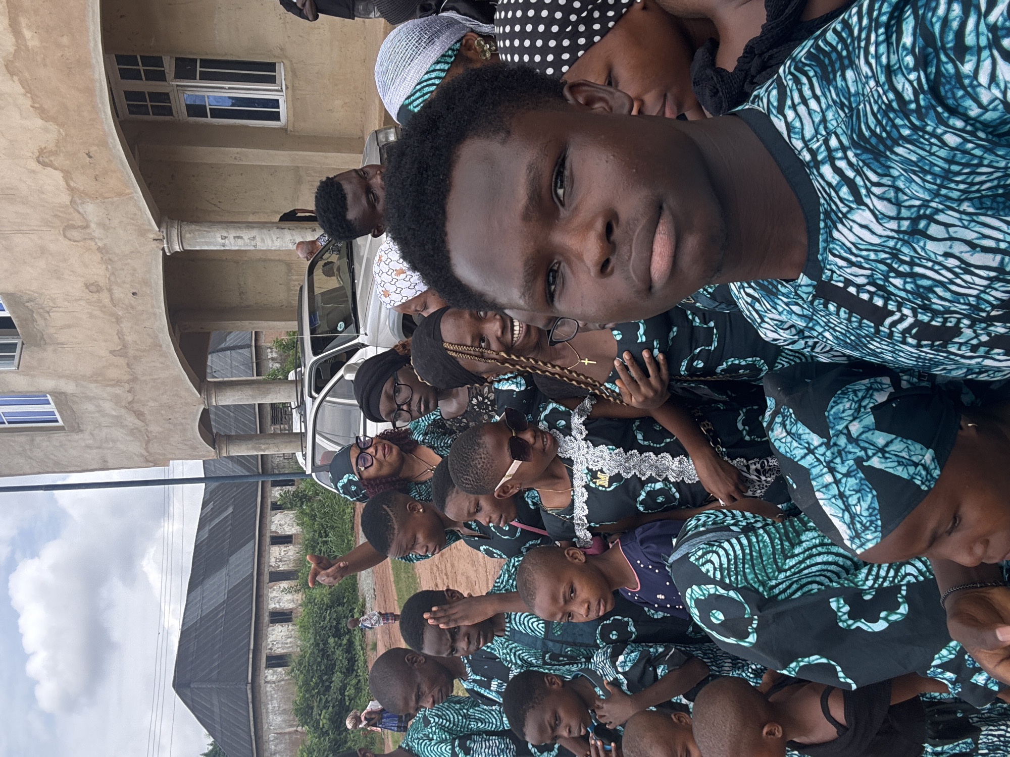 A group of people are gathered outside in Eziudo, Nigeria. Most of them are wearing clothing with the same blue and black pattern. They appear to be posing for a group picture. In the background is a multi-story building with several windows, next to a white car. A few other people can be seen standing nearby.