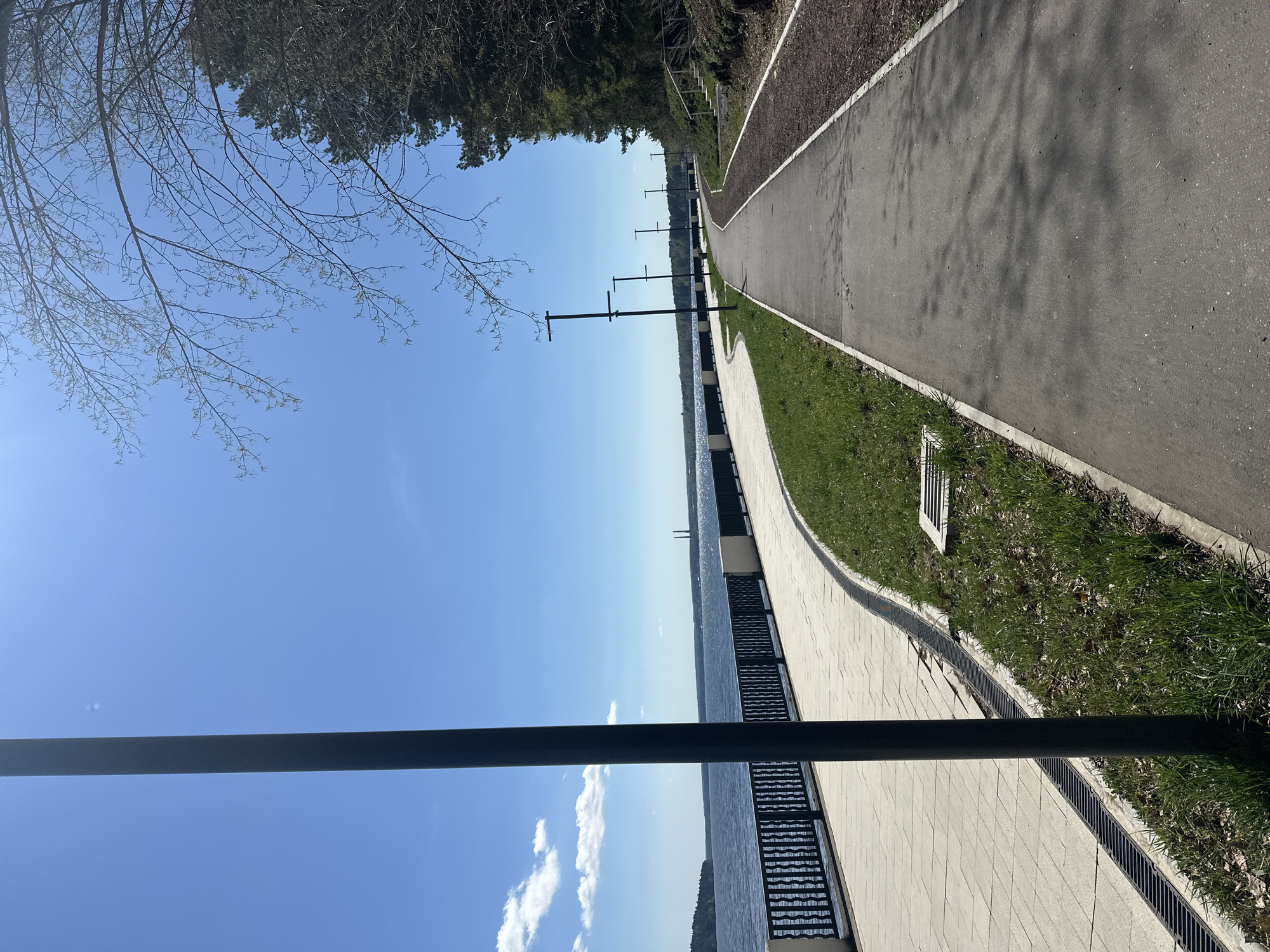 The image depicts a waterfront promenade in Chaikovsky, Russia. A paved walkway runs along the edge of a body of water, with a grassy strip and a further paved area on the land side. A series of modern lighting poles stand along the promenade. A metal railing separates the walkway from the water. The sky is blue with scattered clouds. Trees are visible in the background. The image appears to be taken in spring, judging by the budding trees.
