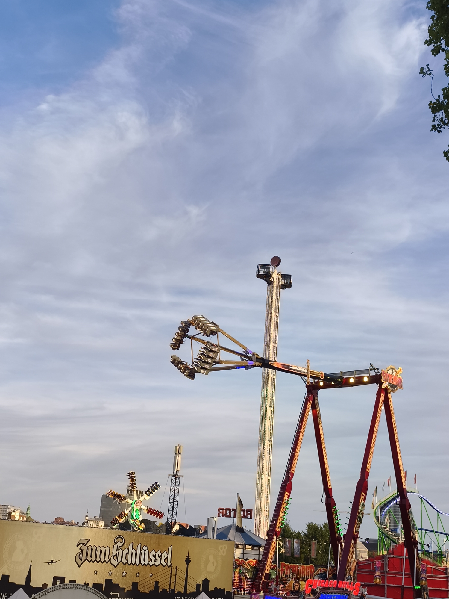 The image shows a daytime scene at an amusement park in Düsseldorf, Germany, likely during the annual Rhein-Kirmes fair. Dominating the foreground is a large red and yellow amusement ride with a central structure supporting two large arms that swing riders. In the background, several other rides are visible, including a tall, slender tower ride and a green roller coaster. A large banner with the text "Zum Schlüssel" and images of Düsseldorf landmarks is also prominent. The sky is partly cloudy. No people are clearly discernible in the image.