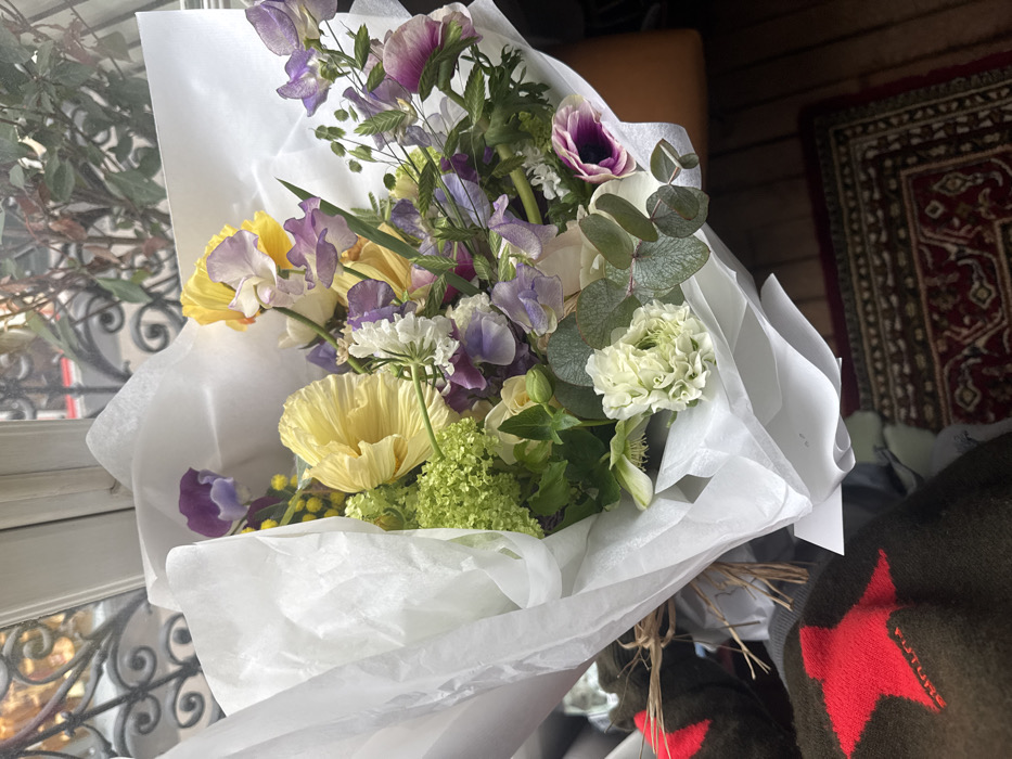 The image displays a vibrant, fresh cut flower bouquet, wrapped in crinkled white tissue paper and secured with natural twine. The bouquet contains a variety of blossoms, including large pale yellow poppies, purple and light purple sweet peas, white clustered blooms, green hydrangeas, pale roses, deep purple anemones, and eucalyptus foliage. This arrangement is positioned indoors, near a window, through which an ornate black wrought-iron railing is visible, strongly indicative of Parisian architecture. Beyond the railing, blurred green foliage and hints of an urban street scene are discernible. In the lower left portion of the frame, a dark garment, featuring a prominent red five-pointed star and the embroidered word "FUTURE," is present. A segment of a dark red patterned rug on a wooden floor occupies the lower right.