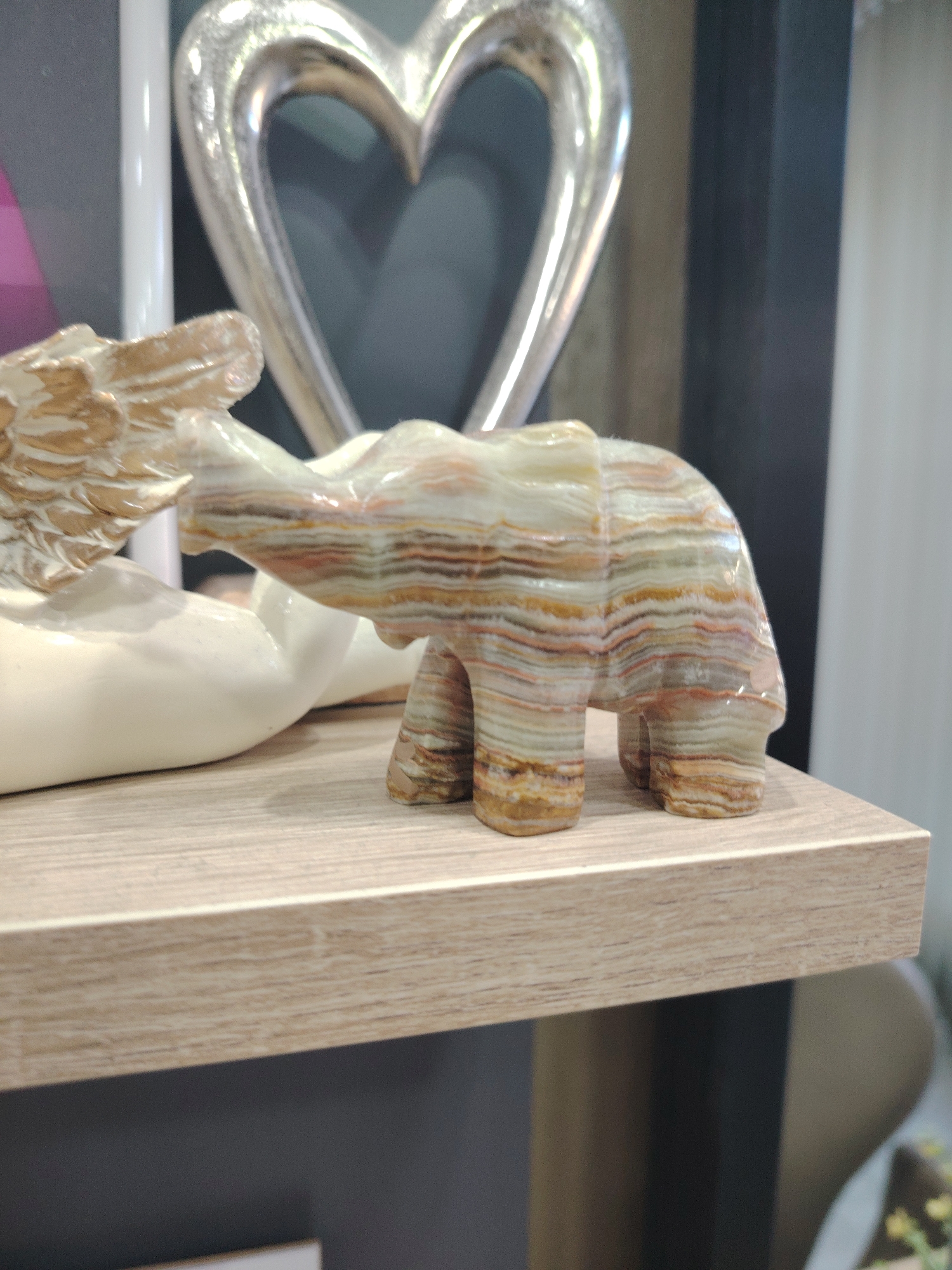 The image displays a close-up, eye-level shot of a decorative, multi-colored stone elephant figurine. The elephant is positioned on a light-colored wooden shelf, angled slightly to the right. The figurine is carved from what appears to be onyx or a similar banded stone, exhibiting layers of white, beige, brown, and reddish-pink. Its trunk is raised, and its four legs are planted firmly.
To the left of the elephant, partially visible, is a light-colored, abstract sculpture, possibly representing a hand or wing, with a textured golden surface. In the background, a large, metallic heart-shaped sculpture with a polished finish is prominent. Behind the heart, the background is out of focus, showing varying shades of gray and dark tones, suggesting an indoor setting. A vertical white element, perhaps a pillar or frame, is also present in the background to the left of the heart sculpture. The lighting appears even and diffused, indicating an indoor environment, possibly a home or shop display. No people are visible in the image. There is no visible text. The overall impression is one of decorative display or collectible items. The location context of Haskovo, Bulgaria, does not provide any specific visual cues within the image itself.