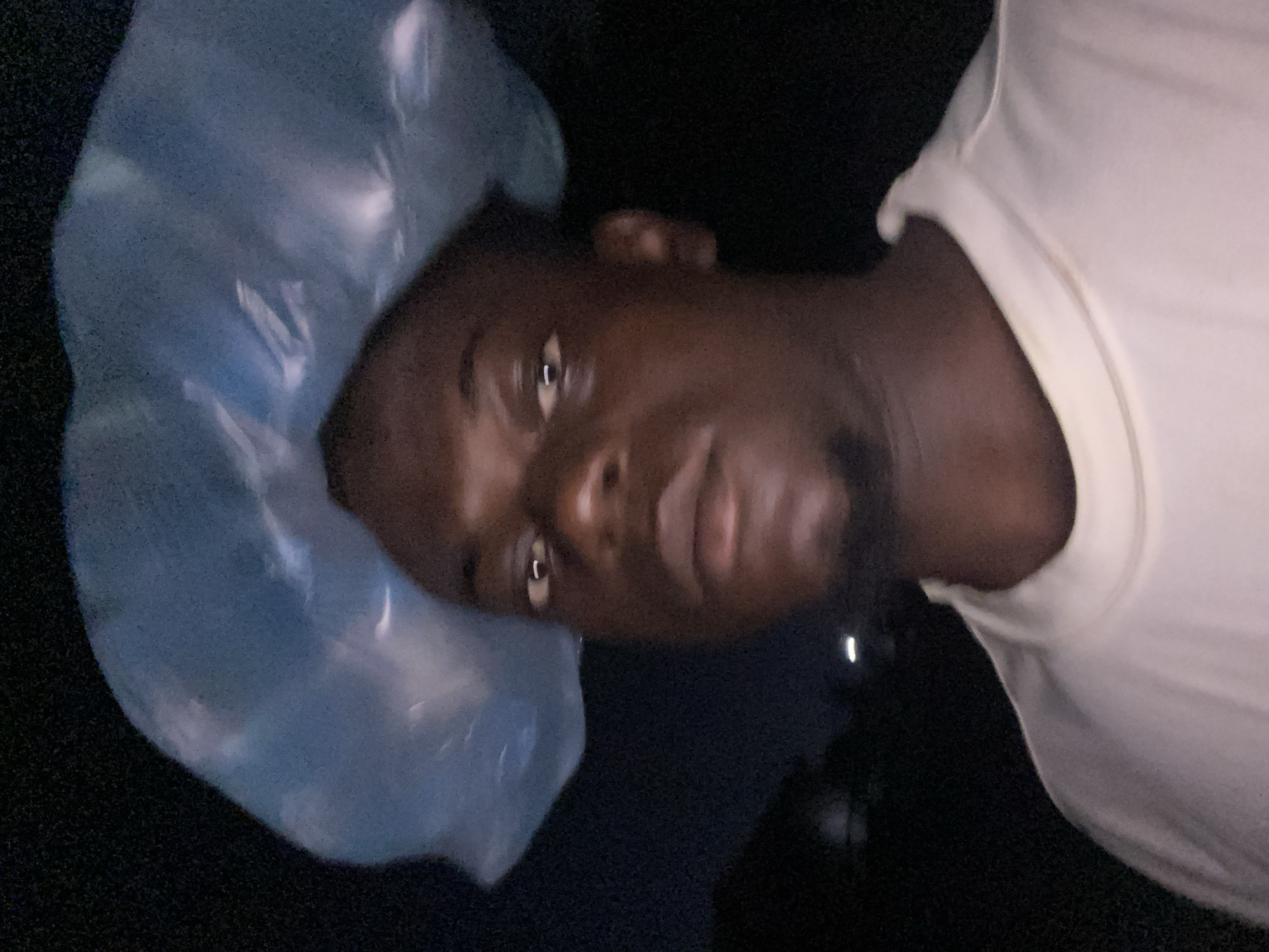 The image is a portrait, rotated 90 degrees counter-clockwise, featuring a person with dark skin looking directly into the camera. The subject, who appears to be male, is wearing a white t-shirt. On their head, covering their hair, is a light blue, translucent, crinkled plastic object that resembles a shower cap or a plastic bag. The background is uniformly dark, suggesting the photograph was taken at night or in a dimly lit indoor setting. A prominent light source illuminates the subject's face from the front, creating bright reflections in their eyes and on the surface of the plastic head covering. The person's expression is neutral, and they are posing for the camera. There are no other identifiable objects, activities, location cues, or visible text in the frame.