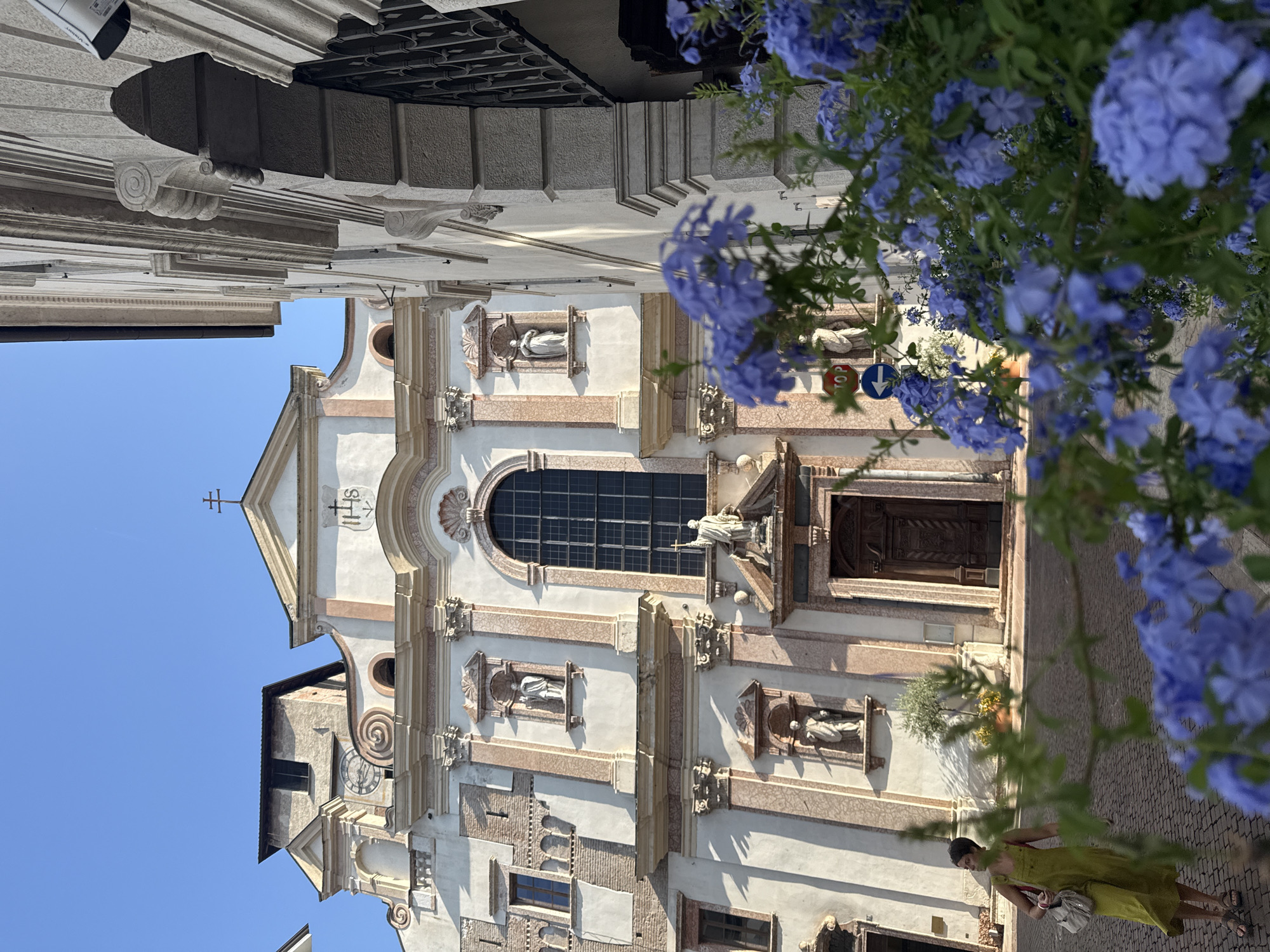 The image shows a view of the Church of Santa Maria Maggiore in Trento, Italy. In the foreground, a woman in a yellow dress walks on a cobblestone street. She is carrying a bag over her shoulder. Blue flowers are in bloom, obscuring part of the scene. Behind the woman and flowers, the white and pink facade of the church is visible, featuring architectural elements like columns, niches with statues, and a large arched window. A cross tops the pediment of the church. To the right, a building with stone detailing and a security camera is visible. The sky is clear and blue.
