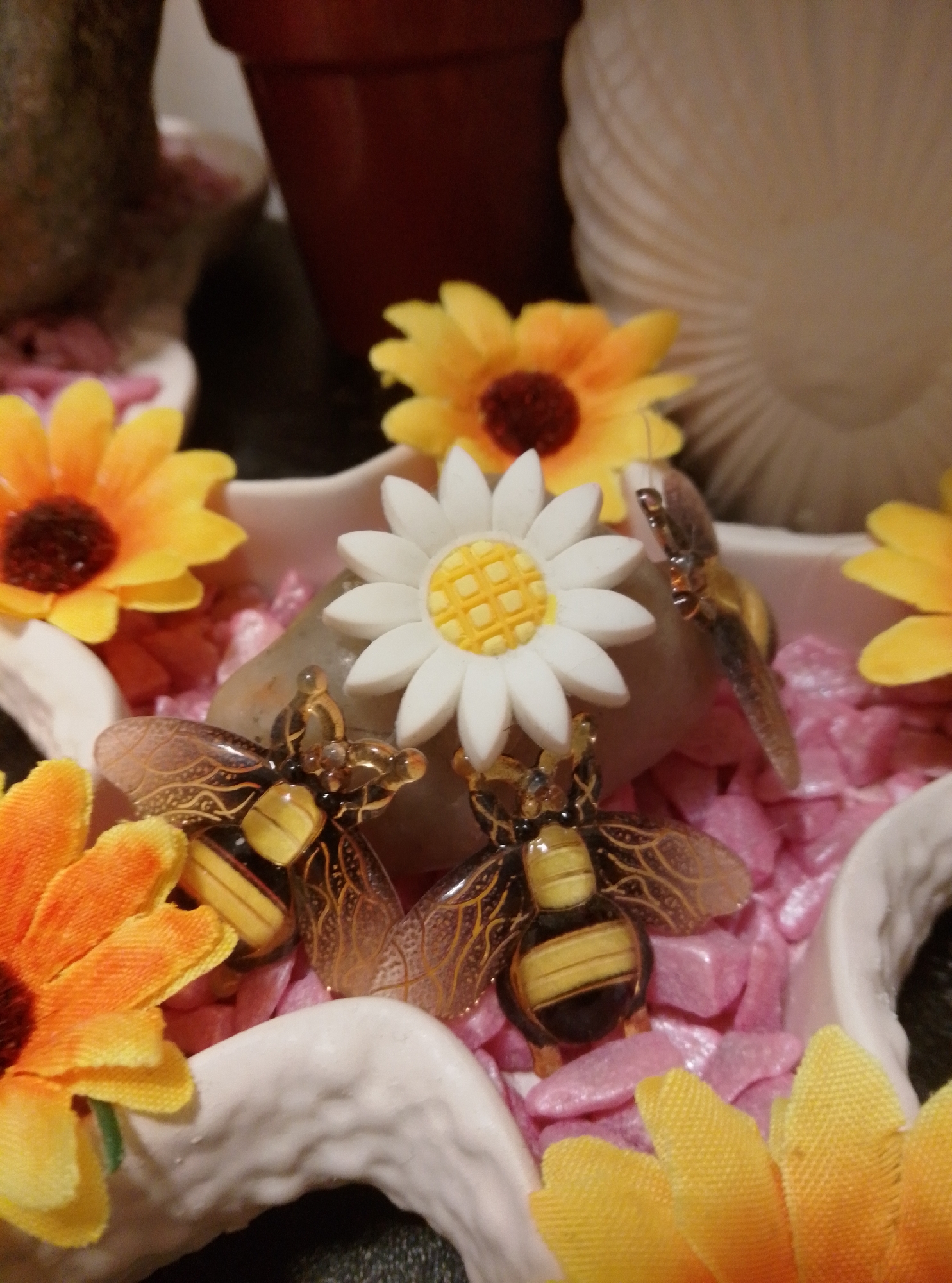 This is a close-up, eye-level shot of a decorative display featuring artificial flowers and two bee figurines. The background is blurred, but suggests an indoor setting with a dark pot and a ribbed, light-colored object. The foreground is dominated by pink, pebble-like material.
In the center of the image, a white artificial daisy with a yellow center is placed. To the left of the daisy, a small, clear or translucent stone is visible.
Two decorative bee figurines are positioned prominently. The bee on the left has transparent wings with dark, intricate patterns, and its body is black and yellow striped with golden accents. The bee on the right is partially obscured but appears similar in design.
Artificial flowers with bright yellow petals and dark centers are visible on the left and upper right sides of the frame. Another artificial flower with orange and yellow petals is on the lower left. The flowers and bee figurines are arranged within a white, textured dish or container with a ruffled edge.
The scene is likely a close-up of a decorative arrangement, possibly a tabletop or shelf display, and there is no indication of a specific location in Wedel, Germany.