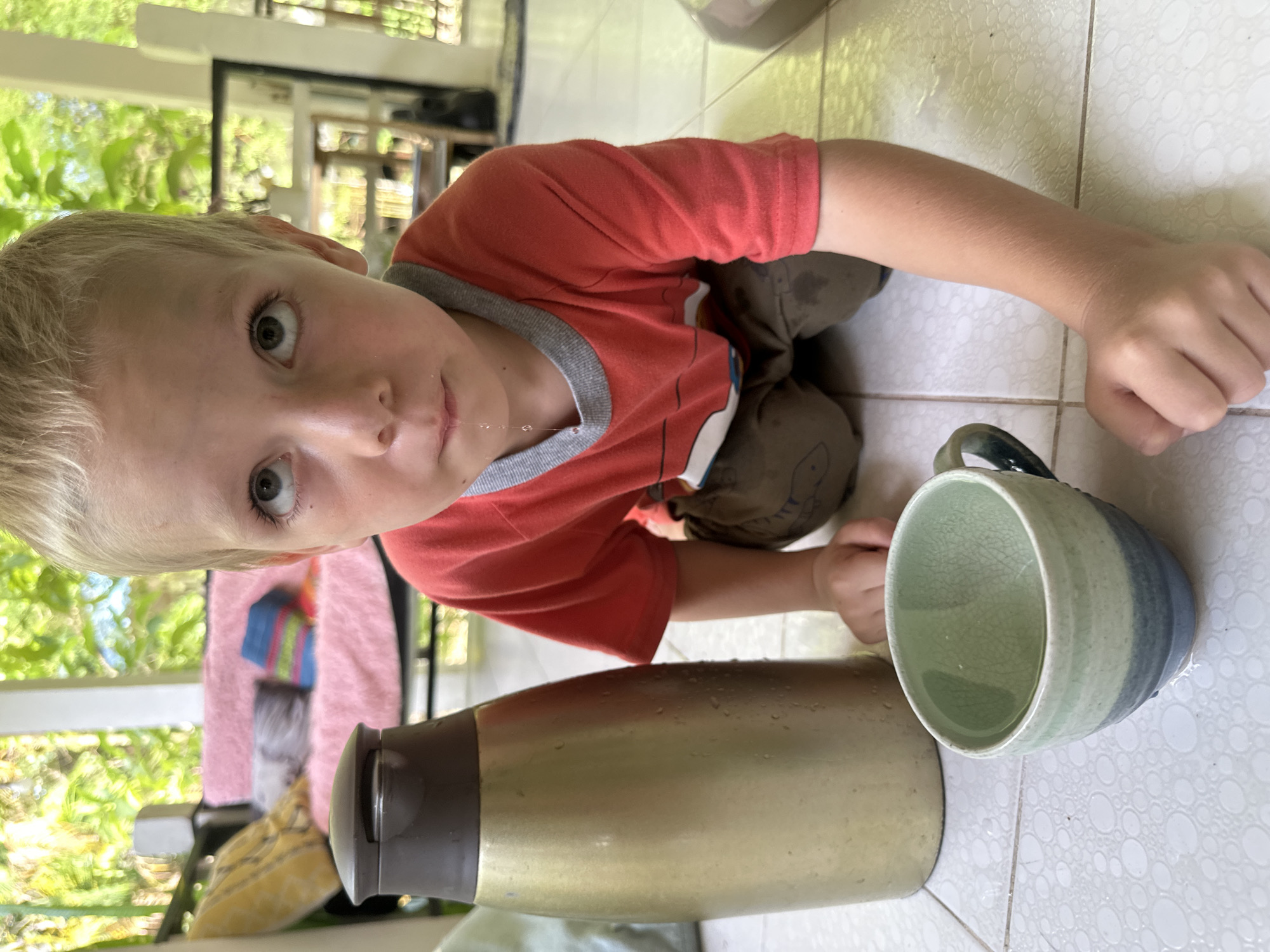 A fair-skinned, blonde-haired boy with light eyes is kneeling on a white tiled floor. He is wearing a red shirt with a gray trim. A string of saliva is extending from his mouth. Next to him on the floor are a gold thermos and a ceramic mug containing liquid. The scene is outdoors, with green foliage visible in the background, suggestive of the location in Baan Chaloklum, Thailand.