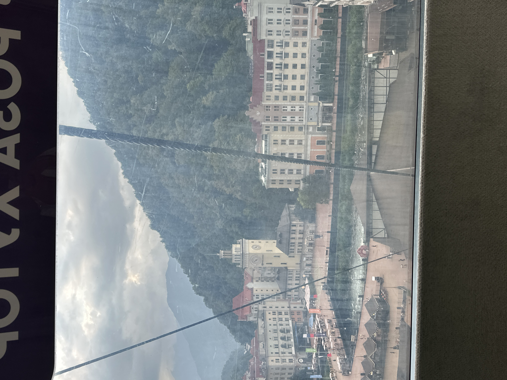 The image displays a view of the ski resort town of Estosadok, Russia, as seen from a cable car or similar elevated vantage point. The scene is dominated by buildings with red roofs, reminiscent of alpine architecture, and a towering clock tower with a spire. Green, tree-covered mountains form a dramatic backdrop to the town. In the foreground, a river or canal flows through the settlement, with a promenade or walkway lining its banks. Small kiosks or market stalls with awnings are visible along the water, and a few people can be seen walking on the promenade. The sky is overcast, with clouds partially obscuring the sun, casting a diffused light over the scene. The image appears to be taken during the daytime.