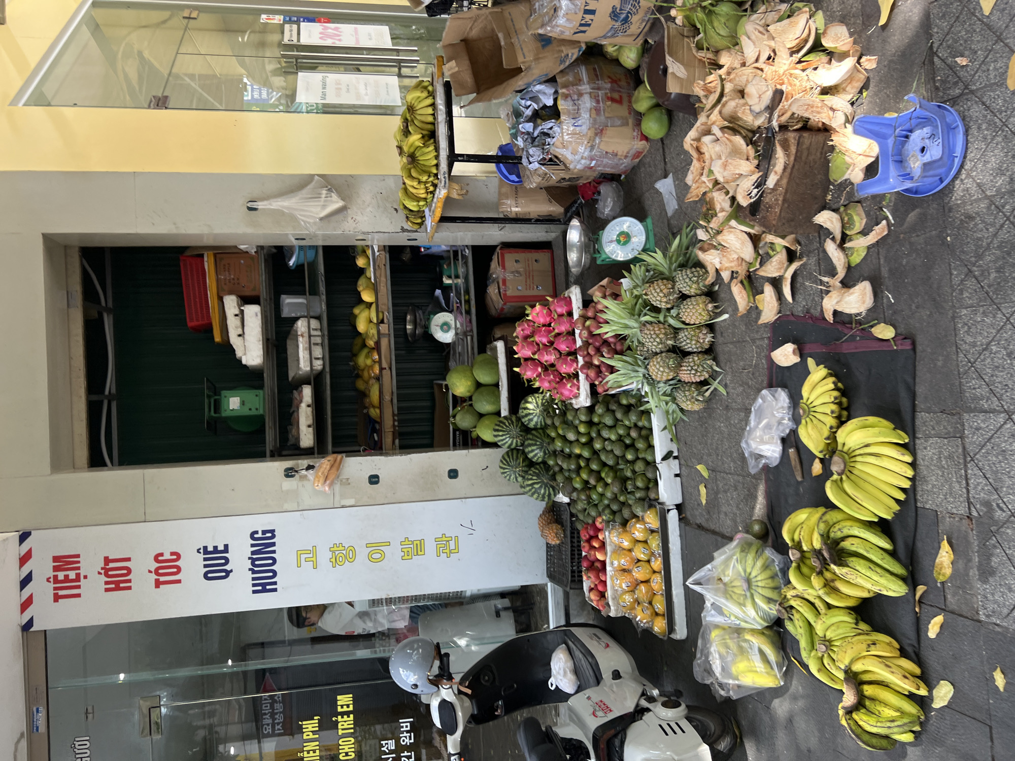 This image shows a fruit stand in Nha Trang, Vietnam. The stand is located on a sidewalk next to a building with a yellow facade and a glass door. A sign in Vietnamese with red and blue lettering hangs vertically on the building. The fruit stand is stocked with a variety of fruits, including bananas, pineapples, dragon fruit, and oranges. There are also some coconuts that have been cut open and discarded. A scale is visible in the background, as well as some boxes and bags. A white scooter is parked on the sidewalk to the left of the fruit stand. A person wearing a white shirt can be seen inside the building. The ground is paved with grey tiles, and there are some fallen leaves scattered around.