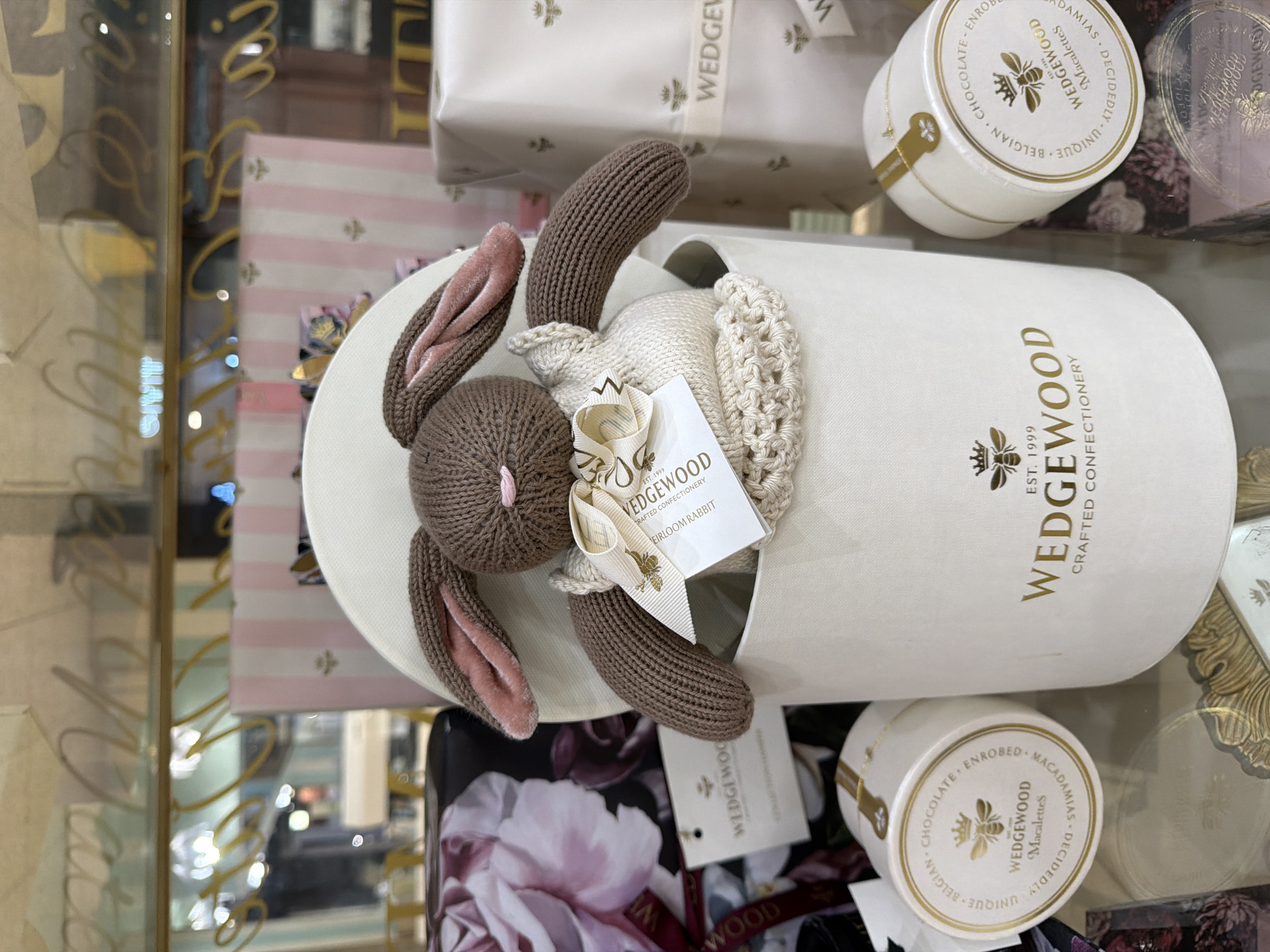 The image shows a display of Wedgwood confectionery items, featuring a knitted bunny inside a cylindrical container with "Wedgwood" printed on it in gold lettering. The container is surrounded by smaller, round boxes of chocolate macadamias, also branded with the Wedgwood logo. In the background, there are glimpses of other merchandise, including a striped gift box, and reflections in what appears to be a display case, suggesting a retail environment. The location, Cape Town, South Africa, is not directly discernible from the visual content.