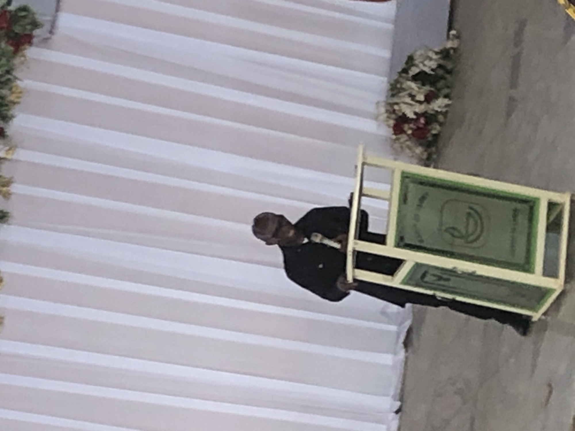 A man in a dark suit stands at a podium with a microphone. The podium is cream-colored and decorated with green panels displaying a symbol. Behind the man, a white pleated curtain hangs and is decorated with floral arrangements. The location is Nteje, Nigeria.