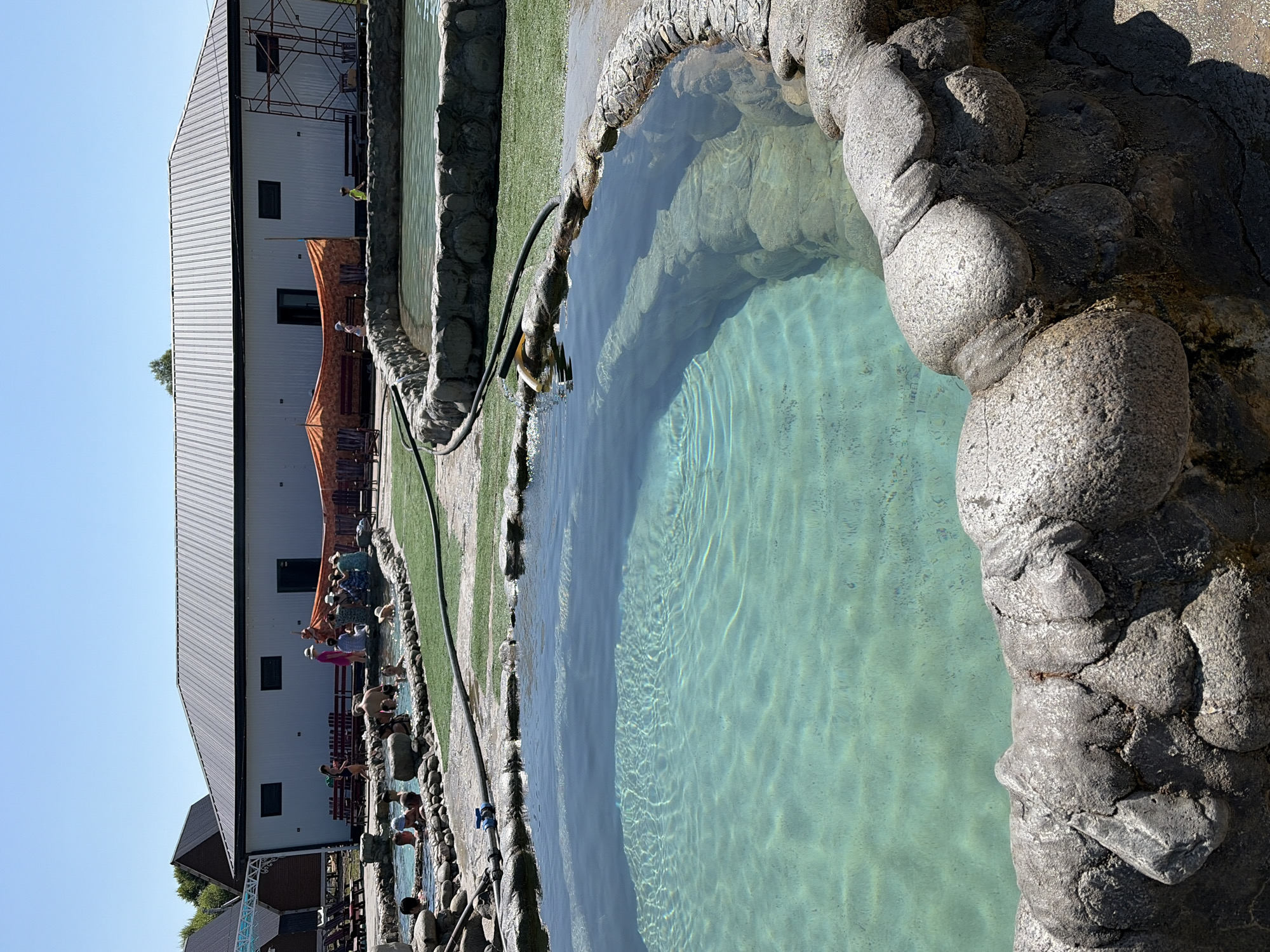 An exterior, eye-level, medium shot captures several naturalistic pools filled with clear, turquoise water, surrounded by rough, grey stone and green astroturf. In the foreground, a large, circular pool with terraced stone steps is visible, its surface rippling under the bright, sunny sky. A black hose with a yellow nozzle extends into the pool from the right. In the background, a long, white building with a dark, corrugated metal roof stands under the blue sky. Orange tarps are draped over benches and awnings in front of the building, and several people are seated or standing around the smaller pools to the left. Some individuals are in the water, while others are on the stone edges. The overall scene depicts a public recreation area with natural-themed water features.