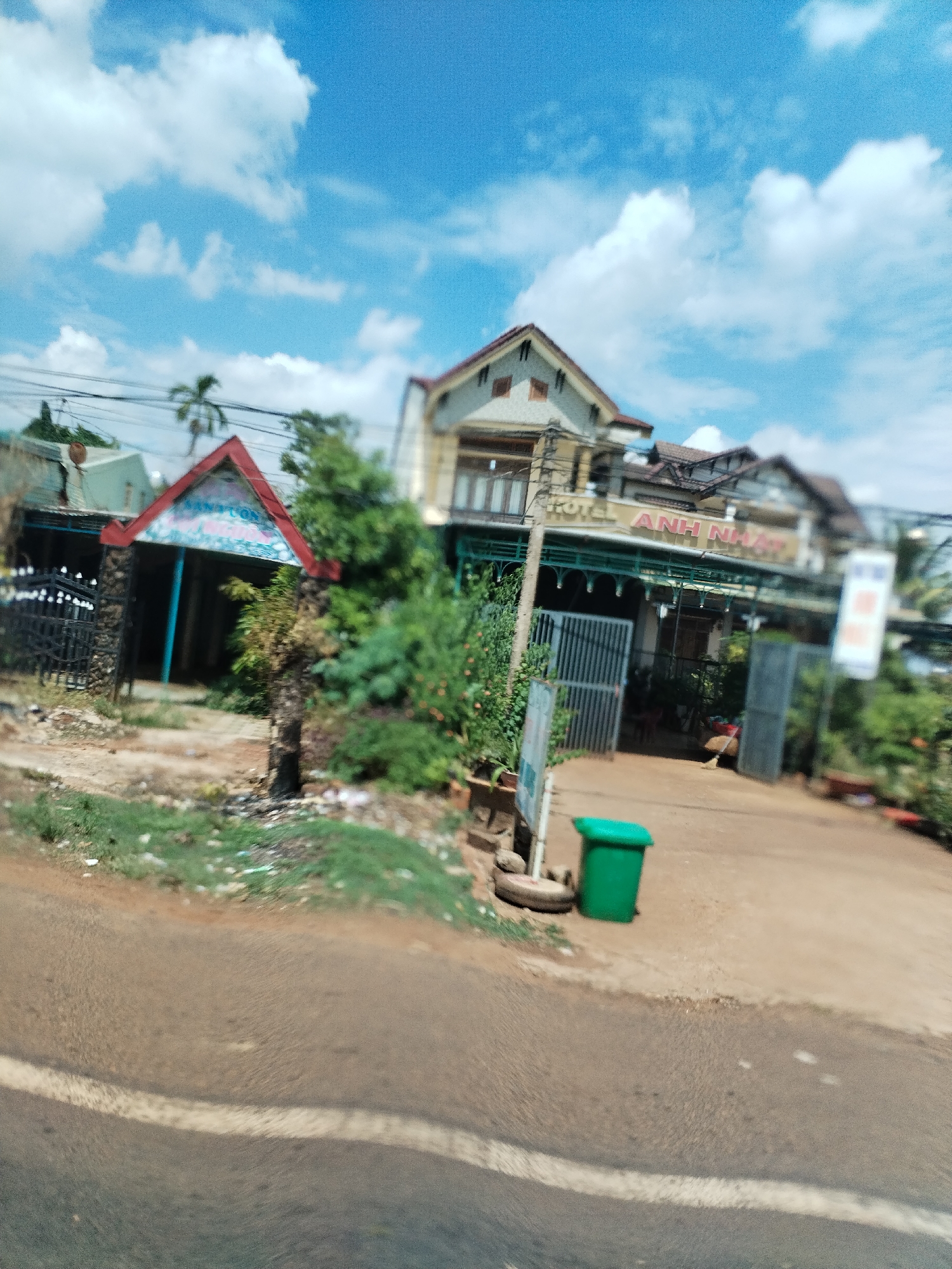 The scene depicts a street in Thôn 4, Vietnam, with buildings on the roadside under a partly cloudy sky. The buildings appear to be commercial establishments, including a "HOTEL ANH NHAT" and another with a sign reading "SAM SON". A green trash can sits on the side of the road. The road surface is visible, with a white line marking a lane. There are no people visible in the image. The environment includes greenery such as trees and shrubs.
