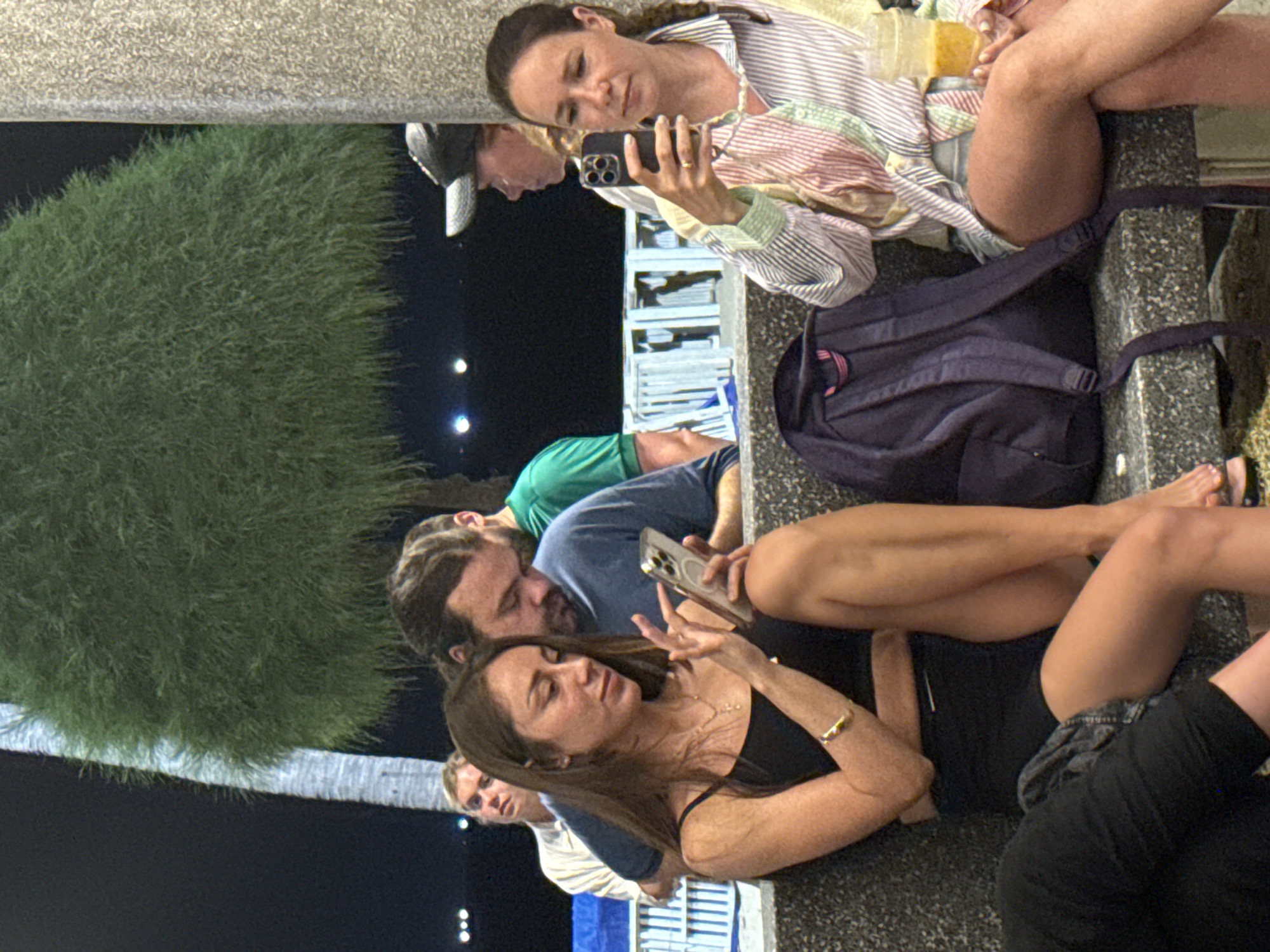 A group of six individuals are gathered outdoors in Nha Trang Ward, Vietnam, at night. A woman in a striped shirt sits cross-legged, looking at her phone. Behind her, a person with a baseball cap looks at the same phone. Next to them is a dark backpack. Below, a woman in a black top holds a phone. A man with long hair looks at the screen over her shoulder. Further to the left, two other people, one in a green shirt and another in a striped shirt, are present, partially visible. A tree is visible next to the group. The background consists of the night sky, lights, and a white structure.