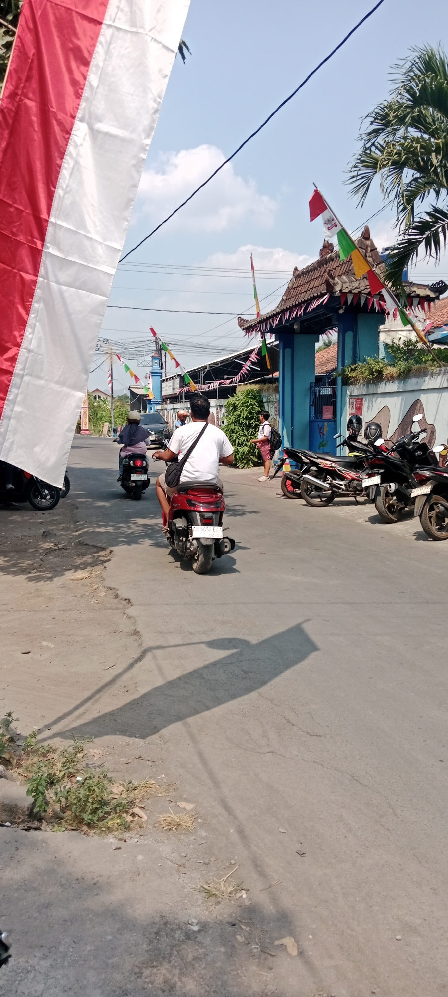 The image captures a street scene in Purwomartani, Indonesia, on a sunny day. In the foreground, a large Indonesian flag, red and white, hangs from the left side of the frame. The street is paved, with some rough patches and sparse vegetation growing along the edges. Several motorcycles are parked on the right side of the street.
Several people are present. A man wearing a white t-shirt and shorts is riding a red scooter, his back to the camera, in the middle of the road. Another person on a scooter is ahead of him, also heading away from the viewer. Further down the street, a person on a scooter with a passenger is visible. On the right side of the street, near a blue building with decorative roof tiles and flags, a young person with a backpack stands.
The blue building appears to be an entrance, possibly to a school or public facility, adorned with colorful pennants. Overhead, power lines crisscross the sky, and palm fronds are visible in the upper right corner. The overall impression is a bustling street scene in a warm, tropical climate, possibly during a national celebration given the prominent display of the Indonesian flag and smaller festive flags.