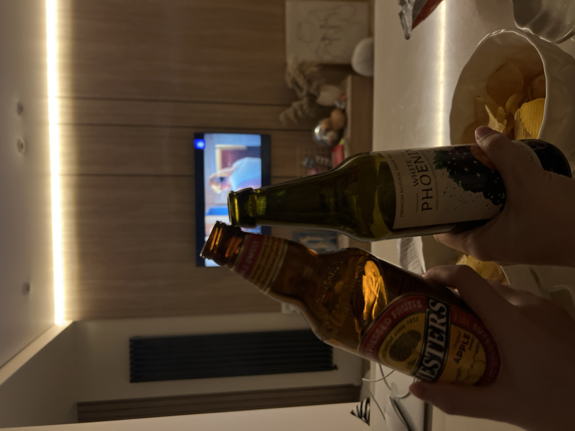 A candid, dimly lit indoor photograph captures two individuals (whose hands are visible) holding bottles as if in a toast, with a television and snacks in the background. In the foreground, two hands hold two different bottles, positioned closely together. The hand on the right holds a dark green bottle with a white label, while the hand on the left holds an amber-colored bottle with a red and yellow label.
The green bottle is labeled "WHITE PHOENIX" with additional text "PREMIUM NATURAL GRAPE" and "HAND-CRAFTED NATURAL GRAPE." The amber bottle prominently displays "ESTER'S" and "APPLE," along with smaller text "PRESSED FRUITS" and likely "SINCE 1888" and "FINEST CIDER."
To the right and slightly behind the bottles, a white crinkled bag or bowl filled with potato chips is visible. In the background, a television screen is on, displaying what appears to be a person or a scene from a show, with a blue icon in the top left corner.
The setting is an indoor space, likely a living room, with warm, dim lighting suggesting evening or night. A vertical LED light strip illuminates a white wall on the left, adjacent to a wooden paneled wall. A dark radiator is visible on a lower wall section. The overall ambiance is relaxed and casual, indicative of a social gathering or a cozy night in.