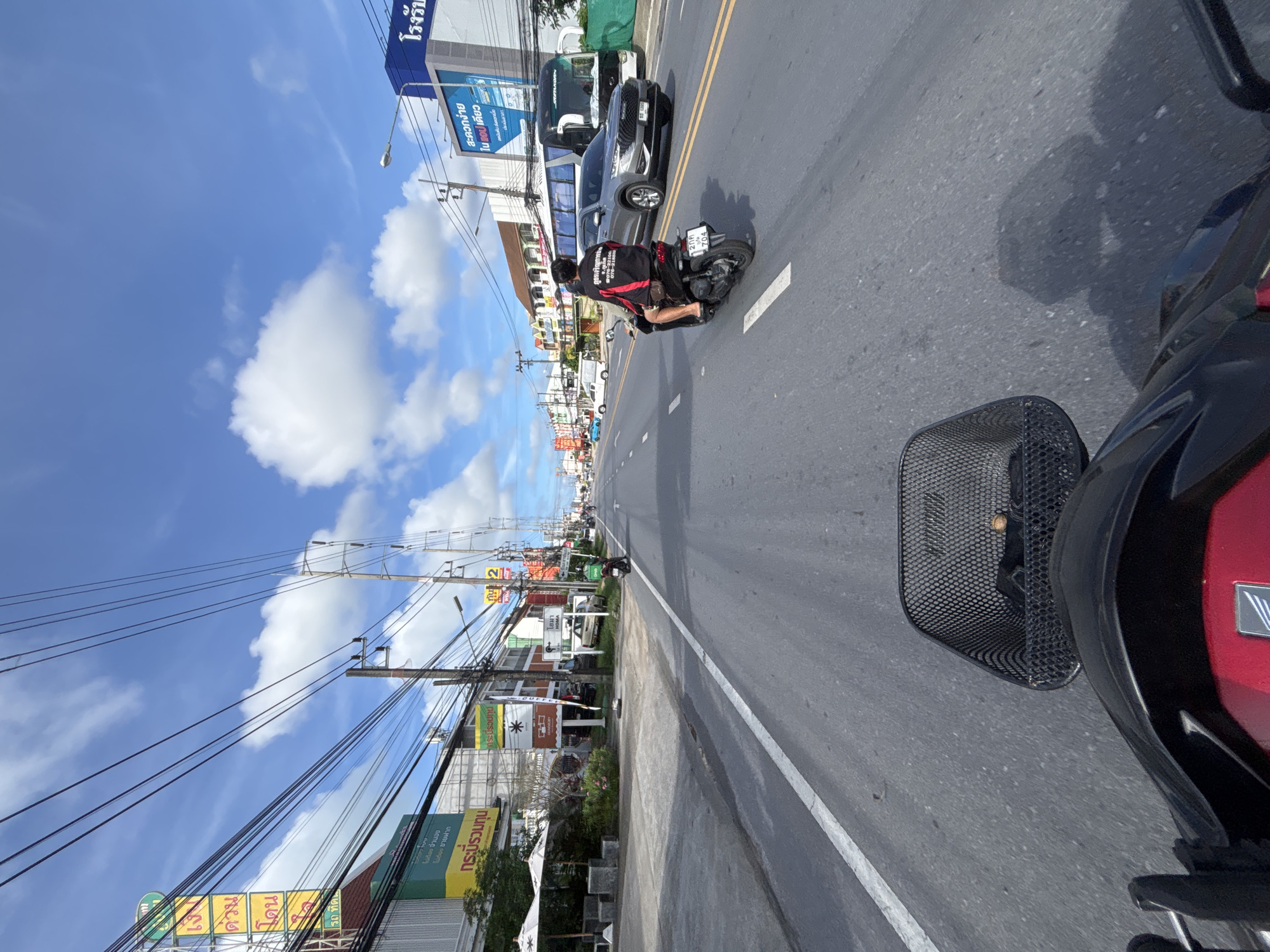 This is a photograph taken from the perspective of someone riding a scooter on a road in Chalong, Thailand. The sky is blue with scattered white clouds. On the left side of the road, there are buildings with signs in Thai script, as well as utility poles with a dense network of wires. On the right side of the road, there is a grey SUV and a white bus. A person on a black scooter is in front of the camera, also traveling on the road. The road is asphalt and has white lane markings. The scooter in the foreground has a black basket attached to its front. The scene depicts a typical urban street environment in Thailand.