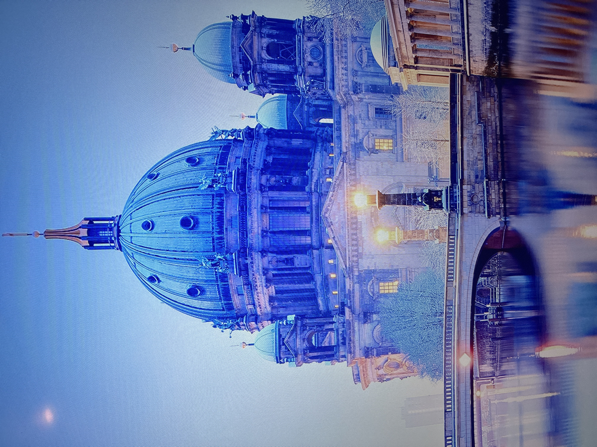 The image displays a grand architectural scene dominated by a large, ornate cathedral under a soft, light blue sky. The main building features a prominent central dome, topped with a slender spire, and is accompanied by smaller domes and towers. Its facade is richly detailed with classical elements, archways, and numerous windows, some of which emit a warm, golden glow, indicating interior illumination.
Adjacent to the cathedral, a substantial stone bridge extends across a body of water, likely a river, reflecting the building's lights and surrounding structures in elongated blurs. Streetlights positioned along the bridge and riverbanks also contribute to the warm lighting accents. Bare-branched trees, suggesting a cooler season such as late autumn or winter, are visible near the cathedral and along the waterfront.
The ambient lighting, a blend of cool blue from the sky and warm yellow from artificial sources, indicates either dusk, night, or early dawn. The scene evokes a sense of historical grandeur and peaceful serenity. There are no people or discernible text visible in the image.
Based on its distinct features, the structure is identified as the Berlin Cathedral (Berliner Dom) in Berlin, Germany, contradicting the provided location context of Sofia, Bulgaria.
