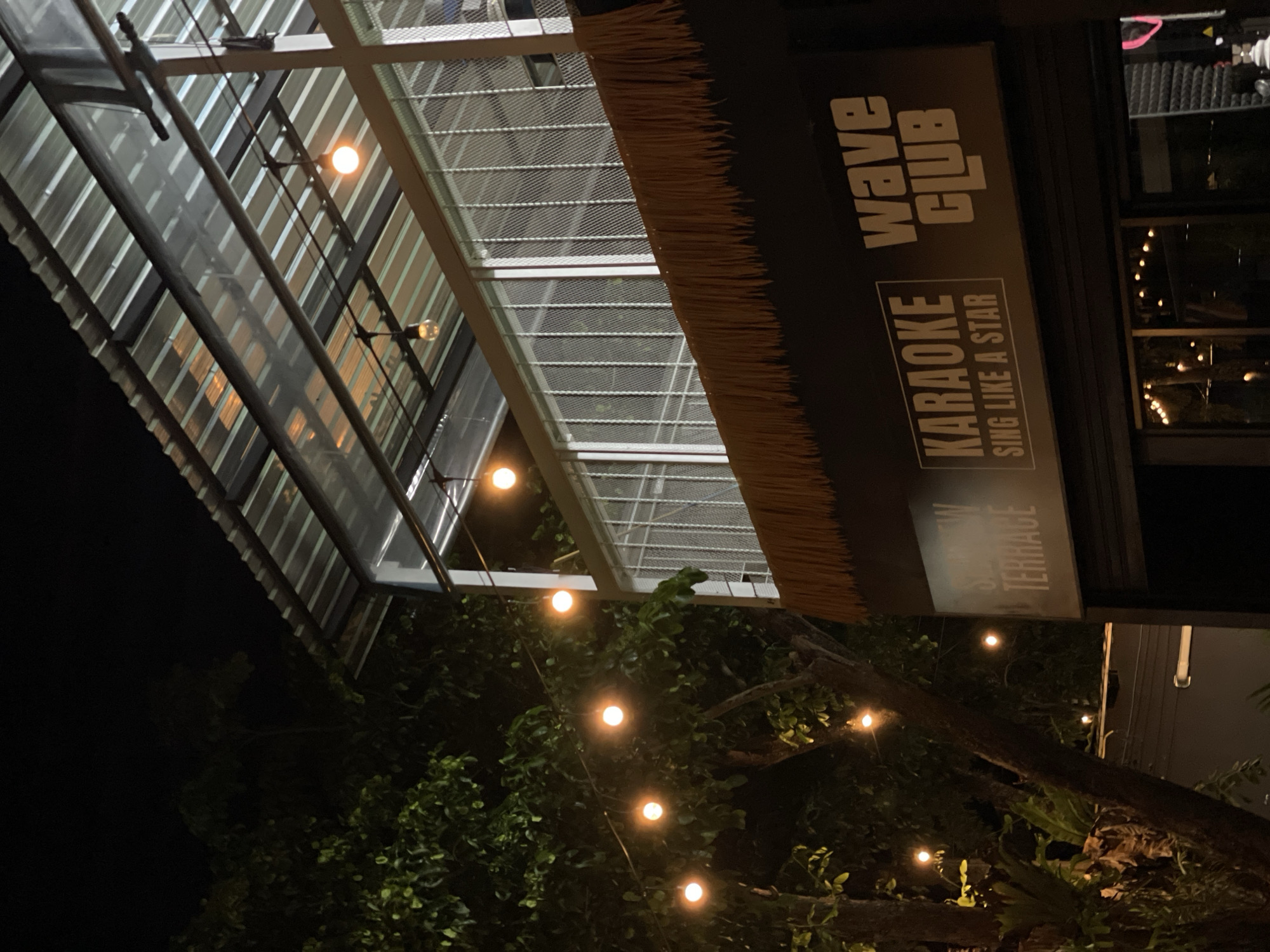 The image depicts the exterior of a karaoke club called "Wave Club" located in Baan Bang Rak, Thailand. The scene is illuminated by strings of warm-toned bulbs strung between trees and across the facade of the building. The building itself features a modern design with a corrugated metal roof and glass-enclosed upper levels with white mesh accents. A thatched-like fringe hangs below the upper levels, directly above the entrance. A sign with the text "KARAKOE SING LIKE A STAR" is visible to the left of the "Wave Club" sign. Trees with lush green foliage frame the left side of the image, adding to the tropical ambiance. The lighting suggests it is nighttime. There are no people visible in the image.