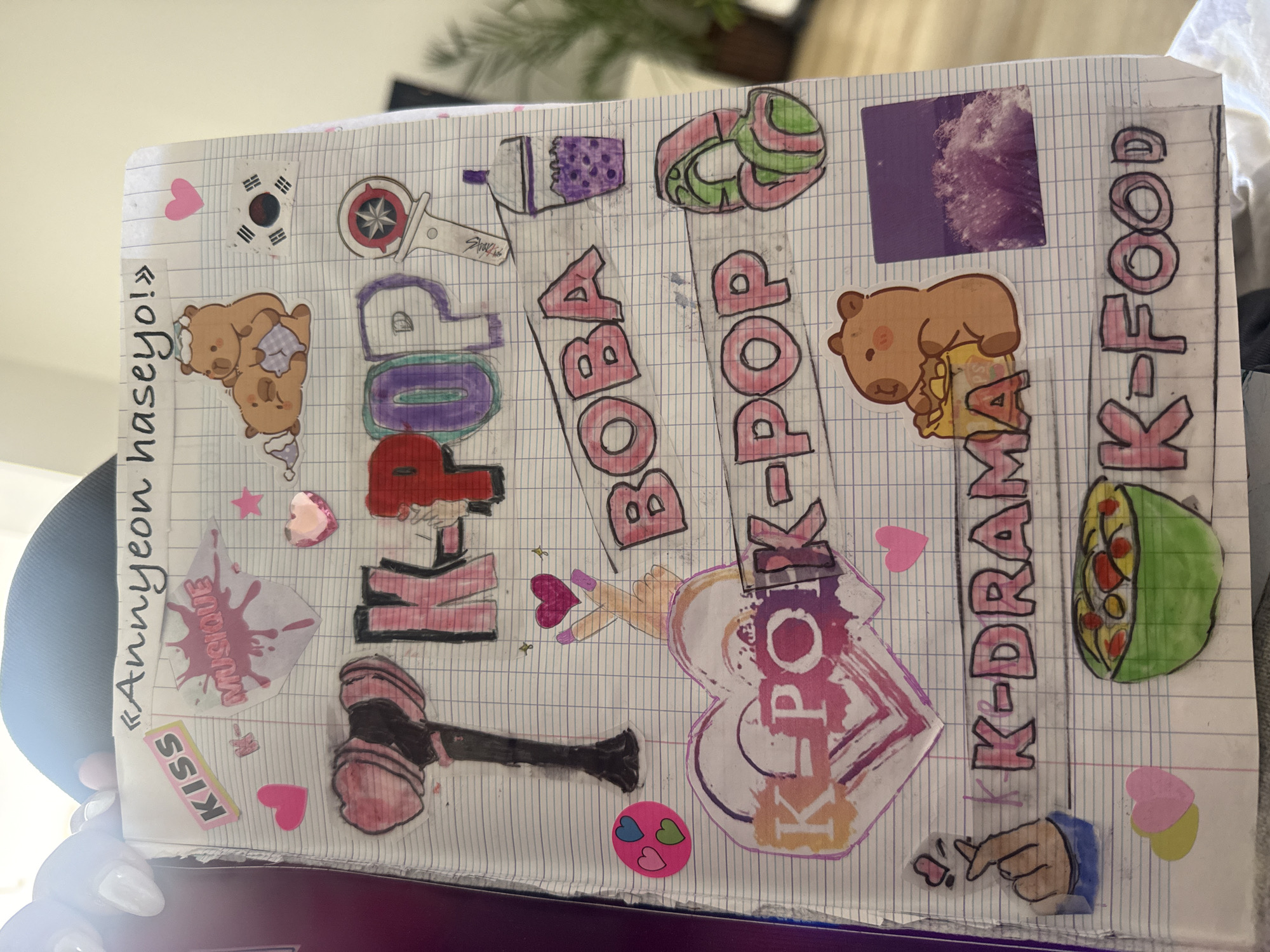 This close-up image displays a notebook page adorned with a vibrant collection of drawings and stickers, largely centered around Korean pop culture and aesthetics. The page features a grid pattern, typical of a school notebook or journal, and is illuminated by bright, possibly natural, light from the left.
Key subjects depicted include various cartoon bears engaged in activities like sleeping or holding items, a boba tea cup, and two K-Pop lightsticks – one clearly labeled "Stray Kids" and another resembling Blackpink's hammer-shaped lightstick. Other drawings include a heart symbol, a hand making a finger heart, and a bowl of food (likely K-Food). Stickers complement the theme, with a South Korean flag, numerous pink and red heart shapes, and a purple square with a textured, cloudy pattern.
The page is heavily decorated with cut-out and drawn text. Visible phrases include "KISS," "Annyeon haseyo!" (a Korean greeting), and "MUSIQUE" (French for "Music"). The dominant themes are highlighted by large, stylized text cut-outs that read "K-POP" (appearing multiple times), "BOBA," "K-DRAMA," and "K-FOOD." The presence of "MUSIQUE" suggests the creator might be French, aligning with the provided location context of Rousset, France.
The overall activity taking place is the creative decoration of a notebook, likely as a form of journaling, scrapbooking, or expressing personal interests. The bright colors and playful designs evoke a sense of enthusiasm and personal expression. The background outside the notebook is softly blurred but suggests an indoor setting with a plant, indicating a casual environment.