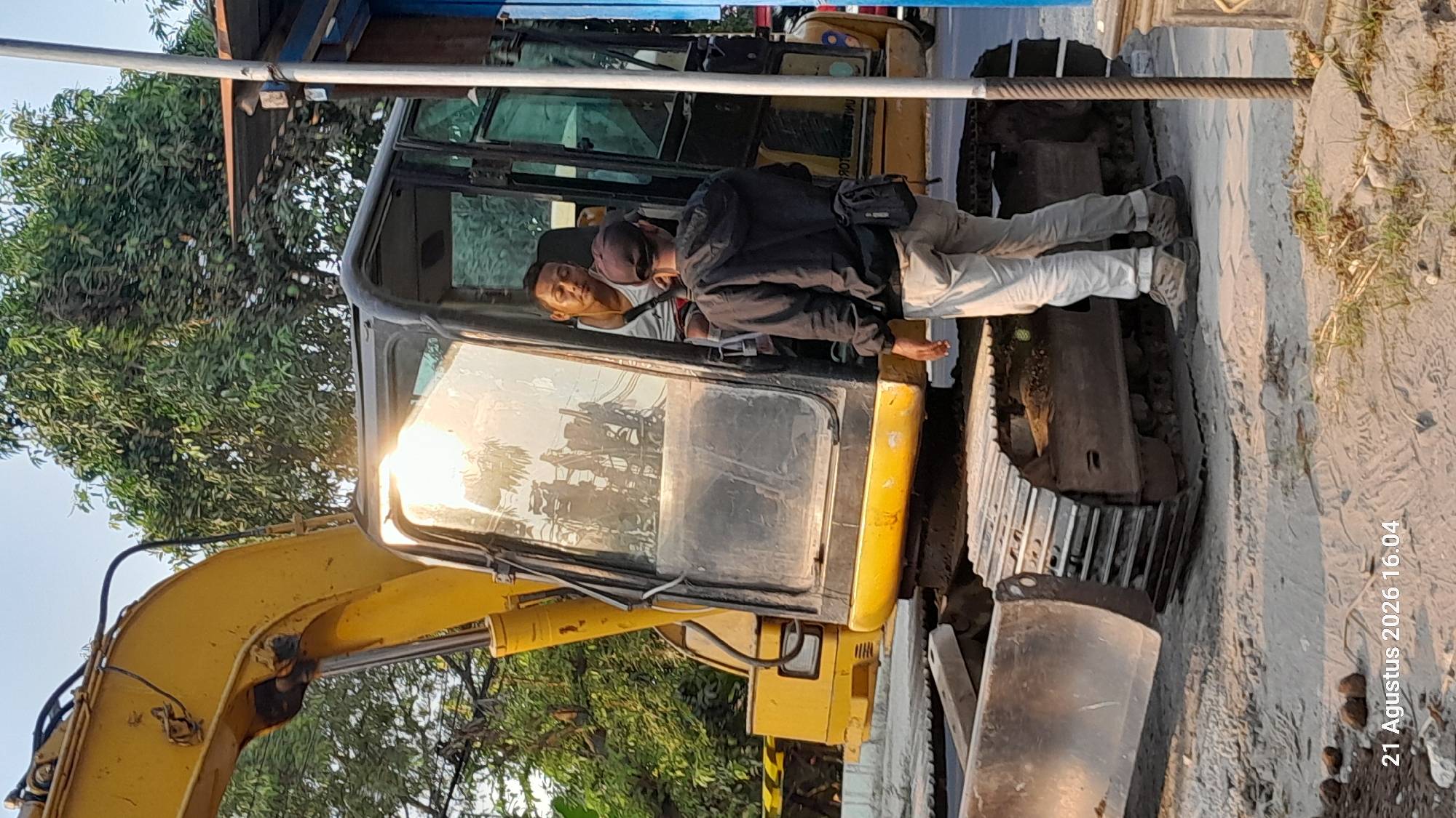 In Takeran, Indonesia, at the timestamp 21 Agustus 2026 16:04, a construction scene unfolds. A yellow excavator dominates the foreground, its tracks resting on the dusty ground. The excavator's cabin, with its large glass windows, reflects the bright sunlight. Inside the cabin, a man wearing a white tank top sits, looking towards another man standing outside. This second man, dressed in a black hoodie and light-colored pants, faces the excavator, his back to the camera. He has a small dark bag slung over his shoulder. The background features lush green trees and a hint of a street or road. The overall environment suggests an outdoor construction or maintenance site in a semi-urban or rural area.