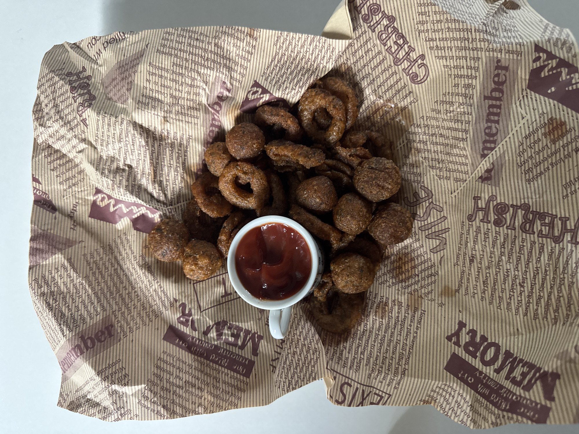The image displays a top-down view of fried snack food served in a brown paper container, set against a plain white background. The food consists of numerous golden-brown pieces, including distinct rings resembling onion rings and smaller, irregular spherical or nugget-like items, suggesting a mix of textures.
Centered within the fried food is a small white cup, similar to an espresso cup, filled with a reddish-brown dipping sauce, most likely ketchup. The cup has a small handle visible on its right side.
The brown paper container is extensively printed with text and decorative elements, giving it the appearance of vintage newspaper or patterned wrapping. Visible English words and phrases include "remember," "cherish," "memory," "filled with," and "home is a warm notion." Other text touches upon themes such as "happiness," "love," "good mood," "patience," and "adversity," suggesting inspirational or sentimental content. The paper also features faint vertical striped patterns in the background of the text.
The scene depicts a casual food presentation, possibly a snack or appetizer, served indoors under bright, even lighting. No people are visible, and the focus is solely on the food and its presentation.
