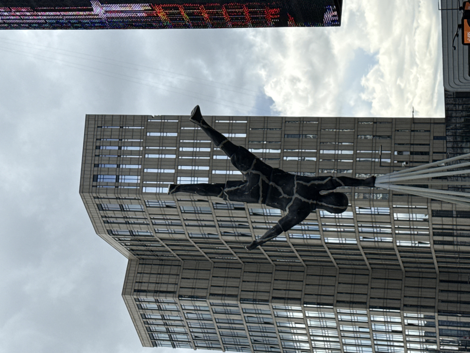 A black, abstract sculpture of a human figure in a handstand is prominently featured in the foreground. The sculpture's surface is textured, resembling cracked stone or bark. It is supported by a white, multi-legged base. Behind the sculpture, a tall, modern building with a grid-like facade of windows dominates the left side of the image. To the right, a portion of a building with a digital display screen showing colorful, abstract patterns is visible. The sky above is overcast with scattered white clouds. The scene appears to be in an urban environment. This sculpture is located in Moscow, Russia.