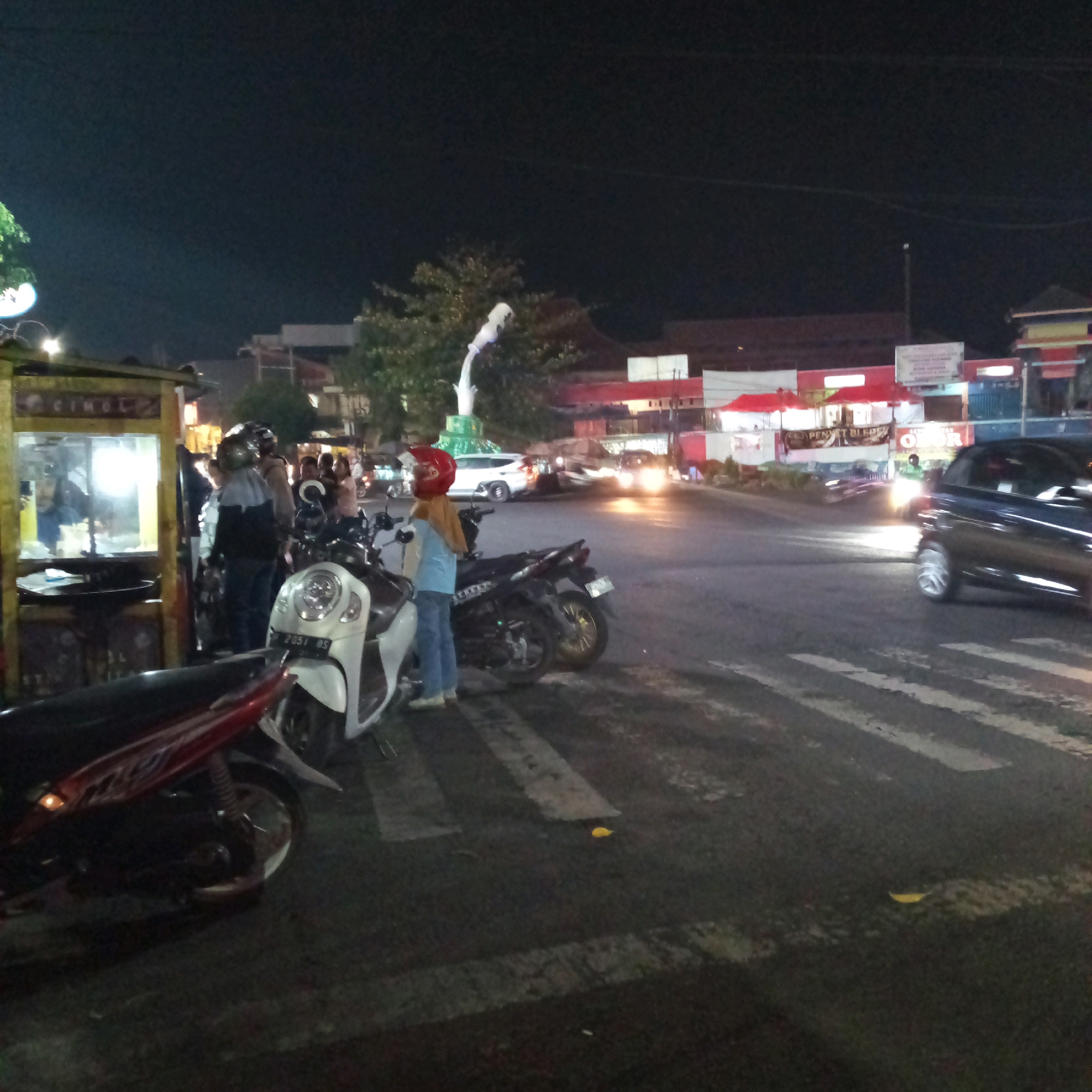 The image depicts a street scene at night in Boyolali, Indonesia, likely near a marketplace or food stall area, given the presence of a food cart on the left. Several motorcycles are parked or being operated on the asphalt road, which has pedestrian crossing markings. In the foreground, a red and black motorcycle is parked. Behind it, a white scooter with license plate "AD 2051 DS" is visible, with a person wearing a blue shirt and red helmet standing next to it. Further back, another motorcycle with a rider wearing a helmet is parked. A food cart with illuminated glass display cases is situated on the left side of the image. In the background, there are buildings with illuminated signs and awnings, typical of commercial establishments. A white statue or monument with a green base is visible in the middle ground. Vehicles, including a dark-colored car on the right with its headlights on, are present on the road, suggesting active traffic. The overall atmosphere is that of a busy evening in an urban Indonesian setting.