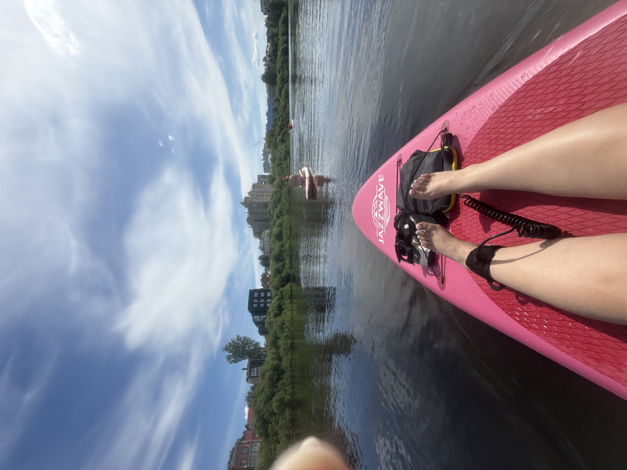 The image presents a first-person perspective from a person on a bright pink paddleboard, floating on a dark, calm body of water, likely the Vologda River in Vologda, Russia. The individual's bare legs are stretched out on the paddleboard, with a black coiled leash attached to the left ankle. A black and yellow dry bag and a pair of white shoes are positioned near the feet on the board, which features the brand "JAZZWAVE."
In the midground, two other individuals on paddleboards are visible. One person, wearing a bikini top and a hat, is sitting on a white paddleboard with their back to the camera, positioned to the right of the center. A third, more distant paddleboarder is partially visible further to the right.
The background depicts the riverbank and the cityscape of Vologda. On the left bank, a dense line of green trees and bushes obscures the lower portions of several buildings. Above the foliage, a multi-story reddish-brown residential building with white windows is visible on the far left. Adjacent to it stands a darker, modern-looking multi-story building. On the right side of the background, beyond more green foliage, a prominent historic architectural complex, strongly resembling the Vologda Kremlin's Bishop's Court, features a large, light-colored building with multiple turrets and arched windows. Further to the right, faint outlines of additional buildings or structures are discernible. The sky above is clear blue with scattered, wispy white clouds, indicating a bright, sunny day.
