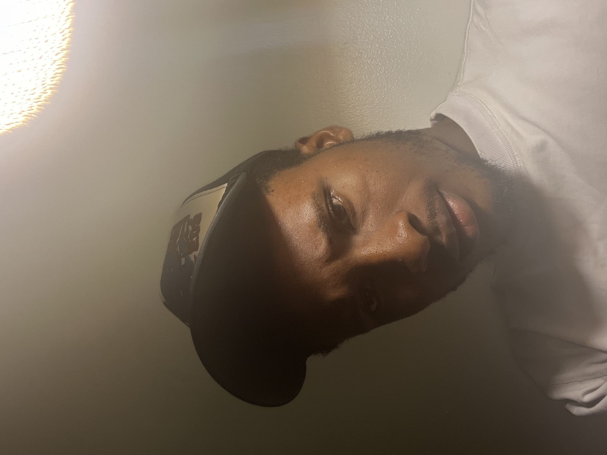 The image captures a close-up, angled view of a dark-skinned male, positioned with his head on the left side of the frame, tilted upwards towards the top-left corner. He appears to be resting or reclining, with a calm and relaxed expression. His eyes are partially visible, looking softly towards the upper right. He has short, dark hair and a beard, and wears a dark cap. A plain white short-sleeved t-shirt is visible on the right side of the frame, covering his shoulder and arm.
A prominent, very bright, warm light source is visible in the upper-left corner, creating a strong lens flare. This intense light illuminates the left side of the man's face (his forehead, cheek, and nose), while the remainder of his face and body are enveloped in deep shadow, creating a dramatic chiaroscuro effect. The background consists of a plain, light-colored, textured wall, suggesting an indoor setting. The overall mood is quiet and contemplative. No visible text is present. This scene is set in Gwarinpa, Nigeria.
