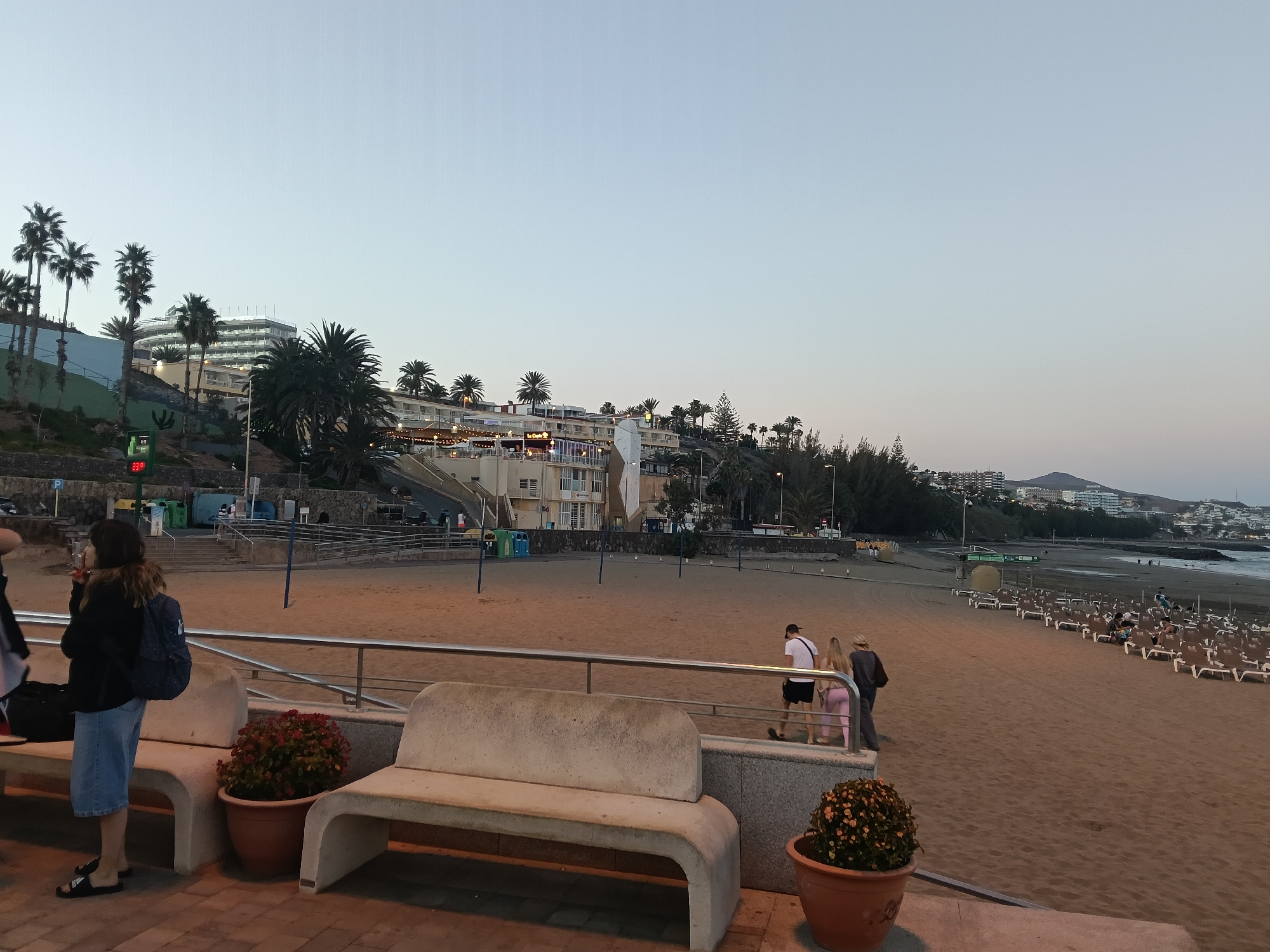 The image shows a view of Playa de las Burras in San Bartolomé de Tirajana, Spain. The sky is dusky, indicating either sunrise or sunset.
In the foreground, there is a raised paved area with modern concrete benches and potted plants. A woman with a backpack is standing near the bench, while three people are walking along the beach path behind the railing.
The beach itself is wide and sandy, with numerous beach chairs arranged in rows. Further out, the ocean is visible.
In the background, there are several buildings and hotels, with palm trees scattered throughout the landscape. The buildings are situated on a slight hill, adding depth to the scene. Streetlights and other infrastructure elements can also be observed.