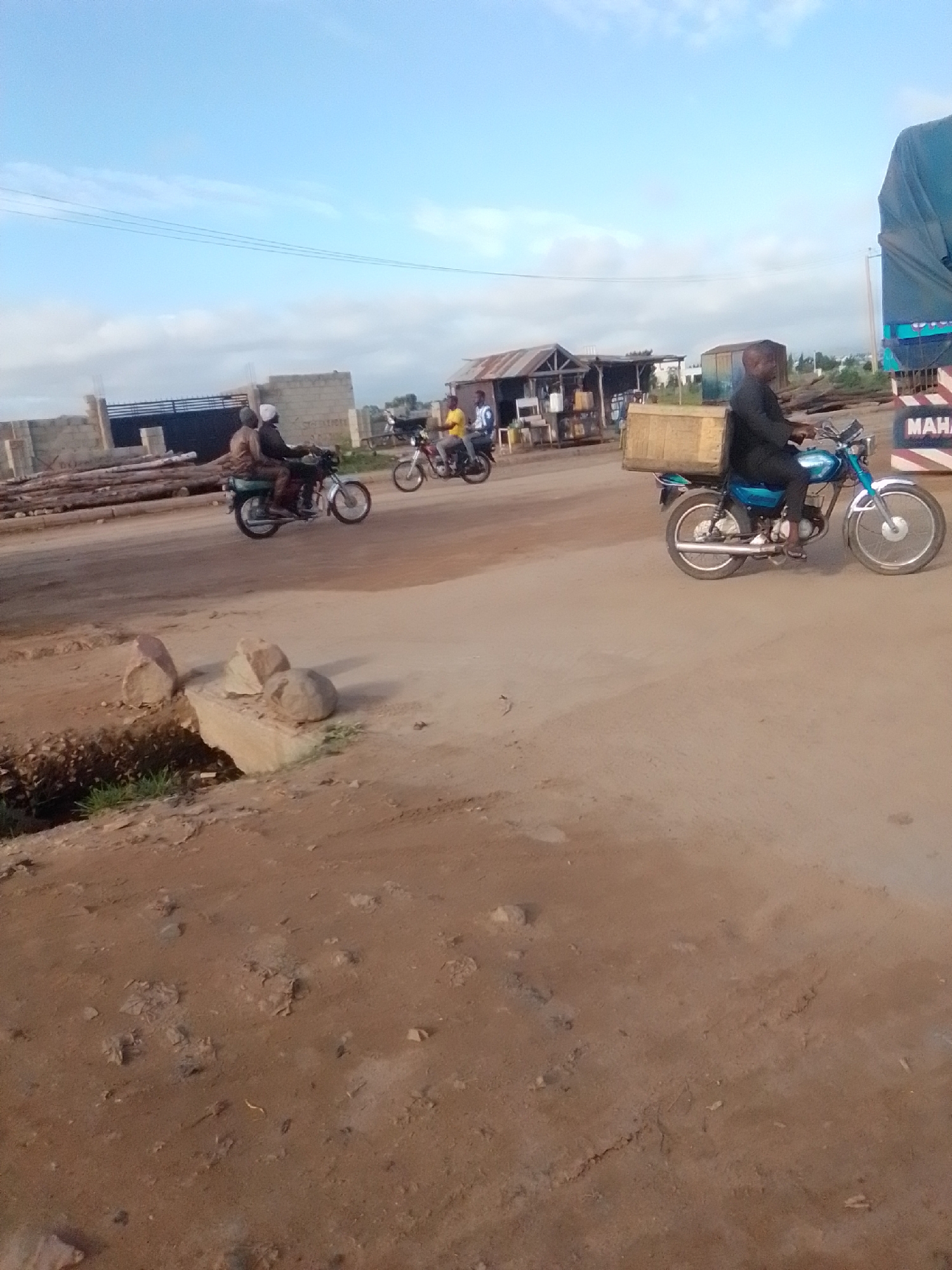 In Kwadon, Nigeria, a scene unfolds with a dusty, unpaved road under a bright, cloud-dusted sky. To the right, a blue motorcycle carries a man wearing a black robe and a wooden box strapped to the back. Further down the road, a blue truck with a tarp is partially visible, displaying the white lettering "MAHA." On the left side of the road, a group of motorcycles are parked. A large pile of logs sits near a partially constructed building with a black metal gate. Another motorcycle with two people is in motion, heading away from the camera, and further in the distance, another motorcycle with two people is also moving. In the foreground, a small ditch with some sparse green vegetation is visible, with several rocks placed around it. The overall environment suggests a working area, possibly a marketplace or construction site, in an open, arid setting.