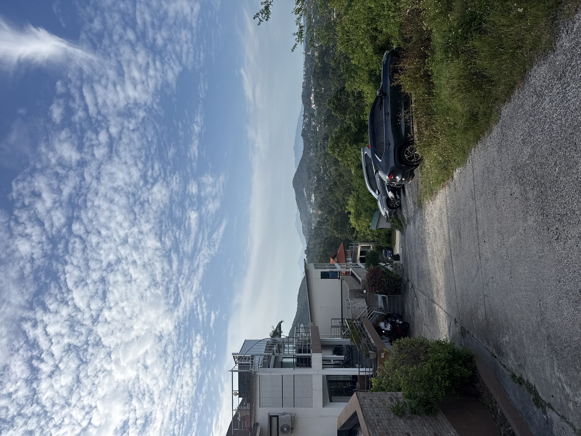 In Ko Samui District, Thailand, a sloping concrete road winds downhill past a modern multi-story white building with balconies and spiral staircases. Adjacent to the building are several parked vehicles, including two SUVs. The road is flanked by lush green vegetation and trees, with more rolling hills and distant mountains visible in the background under a sky filled with scattered white clouds. The scene appears to be a residential area or a resort, given the architecture and the scenic landscape.