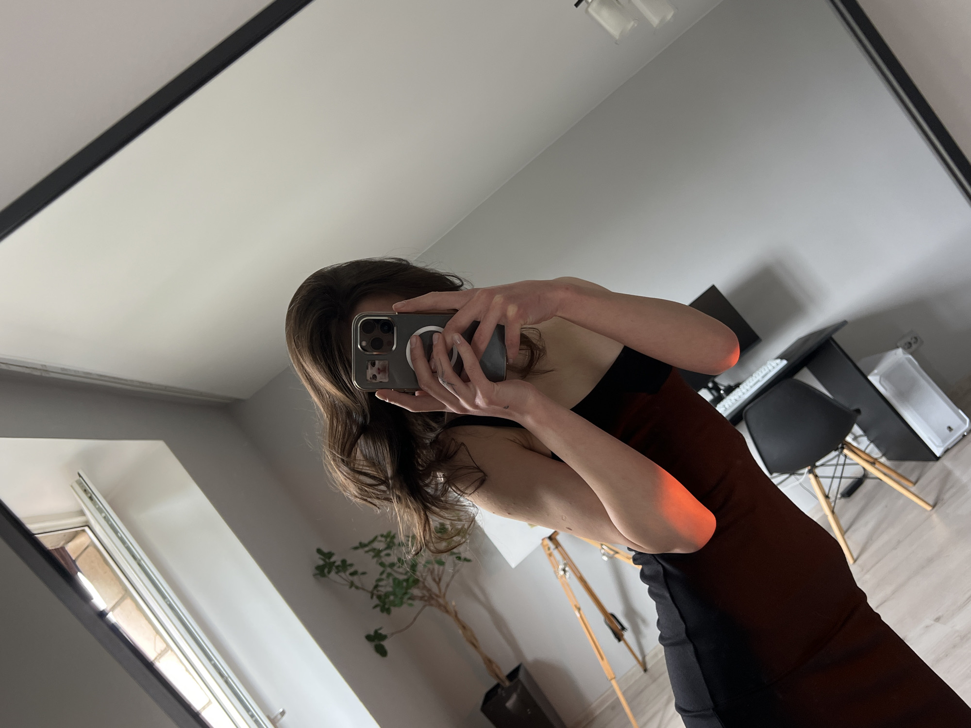 A young woman with long, brown hair is taking a selfie in a mirror. She is wearing a black dress and is holding a phone. The background shows a room with a slanted ceiling, a window, and some furniture, including a plant and a chair. The scene is likely an indoor space in Ternopil, Ukraine.