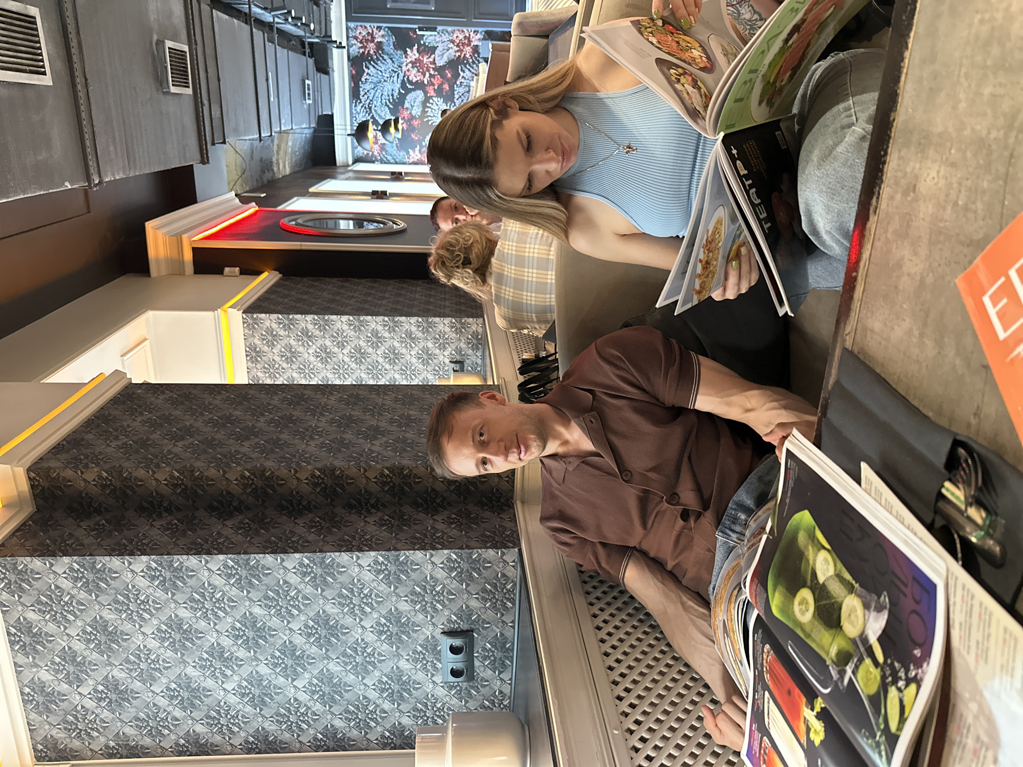In Saint Petersburg, Russia, at what appears to be a cafe or restaurant, a man and a woman are seated on a plush sofa, engrossed in reading magazines. The man, wearing a brown collared shirt, sits to the left, with an open magazine on his lap displaying colorful images of drinks and food. To his right, the woman, in a light blue ribbed tank top and a silver cross necklace, is also focused on a magazine, with another open magazine visible in front of her.
The environment features patterned wallpaper with a repeating floral or geometric design. A white ceramic vase is visible on a ledge to the left, and a dark electrical outlet is mounted on the wall. The seating area has a ribbed texture. In the background, other patrons are visible, including a woman with blonde curly hair and another person. The ceiling has exposed industrial elements and lighting fixtures. A red neon light accentuates a curved architectural feature on the wall. The overall ambiance suggests a modern and somewhat casual dining or lounging establishment.