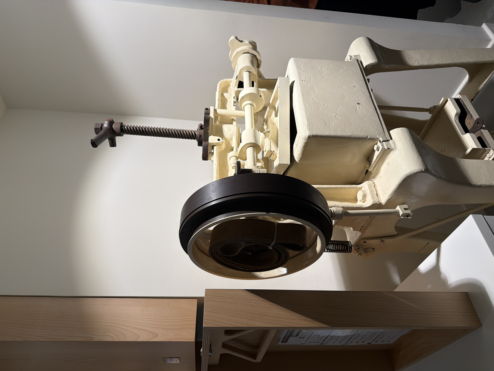 The image shows a vintage-looking industrial machine, painted in an off-white or cream color, likely on display in a museum or exhibit. The machine features a large, dark-colored wheel with a lighter interior ring, beneath which a coiled strap or belt is visible. To the left, a long, threaded metal rod extends from the machine's central mechanism, ending in a T-shaped handle. The machine rests partially above a light wood-toned shelf or cabinet. The background is a simple white wall.
The image was taken at the Musée Würth France Erstein in Illkirch-Graffenstaden, France.