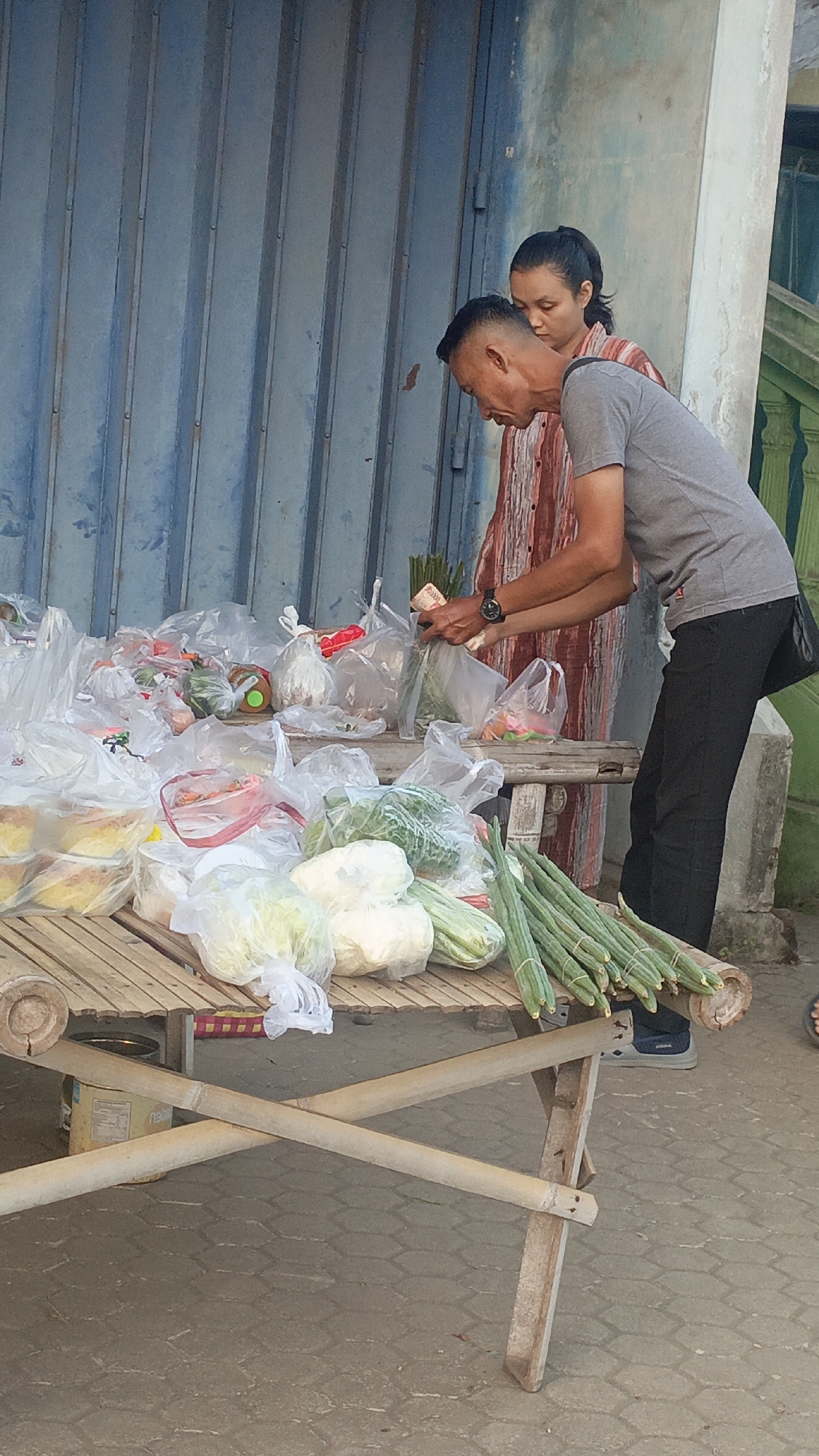 Two individuals, a man and a woman, are standing at a market stall in Srono, Indonesia. The man, wearing a grey t-shirt and black pants, is in the foreground, looking down and handling cash and vegetables. The woman, in a reddish-brown and white striped shirt, stands behind him, her head lowered. They are surrounded by a variety of fresh produce, mostly packaged in plastic bags, on a bamboo table. Some of the visible vegetables include cabbage, cauliflower, and long gourds. The stall is situated in front of a blue corrugated metal shutter, suggesting an outdoor market setting. The ground is paved with hexagonal tiles.