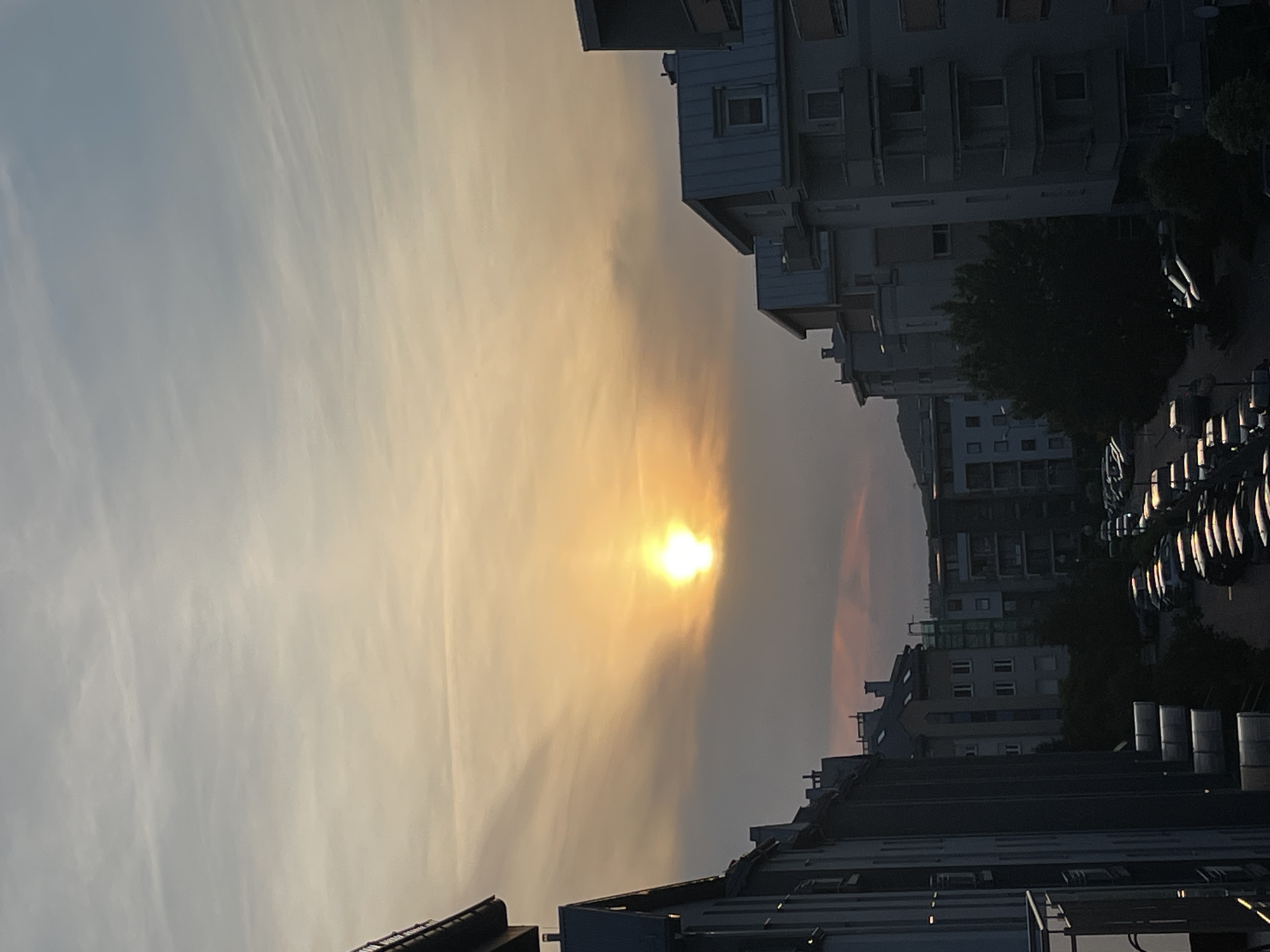 The image captures an urban or suburban scene at either sunrise or sunset, presented with a 90-degree clockwise rotation. The sky dominates the left side of the frame (top if rotated correctly), featuring a striking gradient of colors. A brilliant, glowing sun is positioned prominently in the mid-left, its intense light diffused by a thin layer of haze or wispy clouds, creating a radiant burst of golden-yellow light. The sky transitions from warm yellow and orange hues around the sun to a softer, light blue-grey further away, with subtle cloud patterns visible.
Multiple multi-story buildings are silhouetted against the bright sky on the right side of the image (bottom if rotated correctly), and a smaller group of buildings is visible on the far left (top if rotated correctly). These structures, possibly residential or commercial, show details like windows and balconies but are rendered in dark, muted tones due to the strong backlighting. Dark masses of trees or dense foliage are visible between some of the buildings. A line of parked cars can be seen along a street on the far right, with their reflective surfaces catching some of the ambient light. On some rooftops in the foreground, cylindrical structures, possibly chimneys or vents, are apparent. The scene evokes a calm and atmospheric moment, defined by the low, dramatic light of dawn or dusk. No visible text is present.