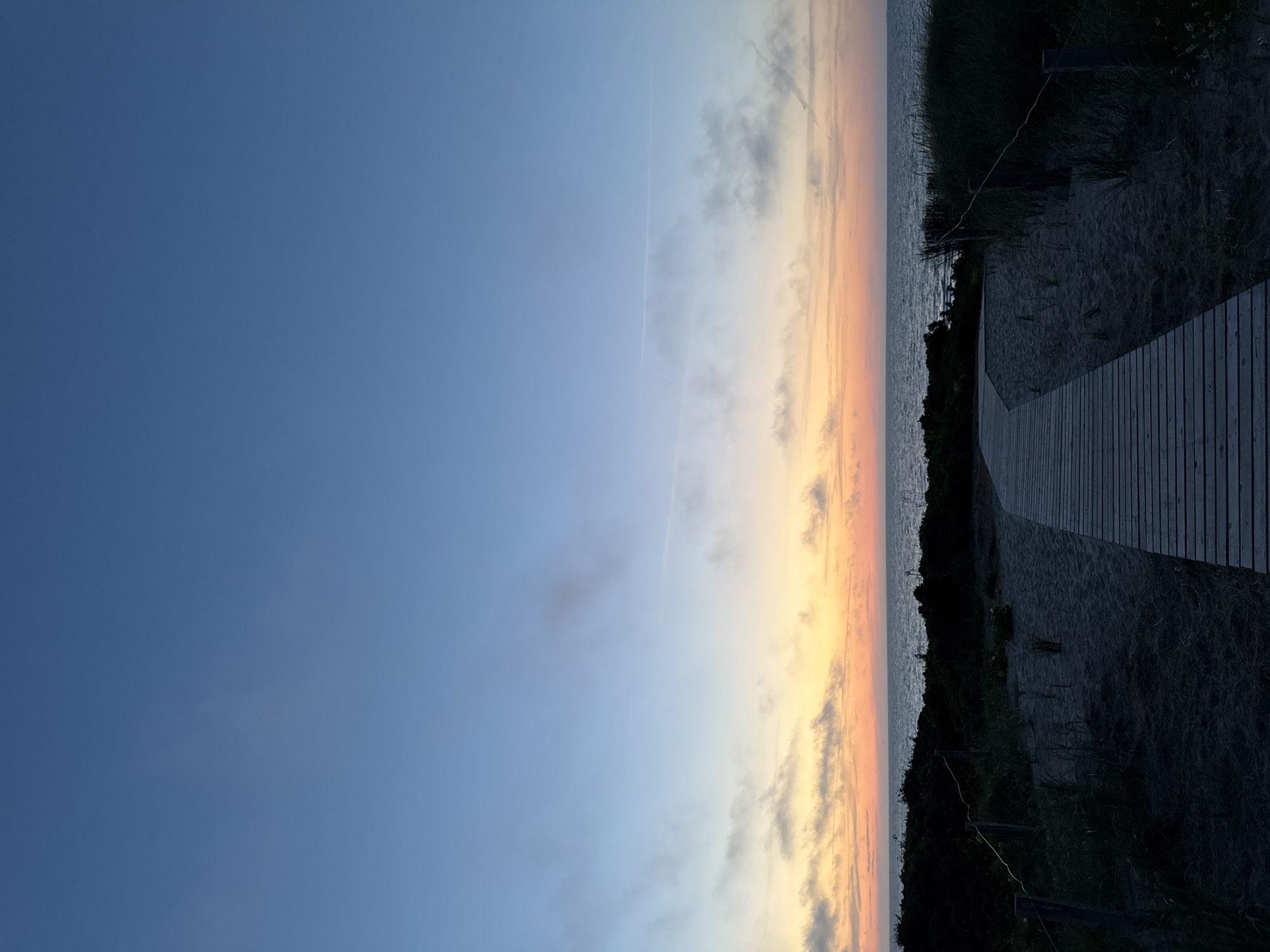 This is a vertical, eye-level, medium shot of a wooden boardwalk leading across a sandy beach towards the ocean at dusk. The sky above is a gradient of deep blue at the top transitioning to lighter blues and then a band of soft orange and pink hues near the horizon, indicating sunset or sunrise. Wispy clouds are scattered across the sky, particularly in the lower portion. The ocean is visible as a calm, dark expanse meeting the colorful sky. The foreground features a dark, sandy beach with sparse vegetation and a wooden boardwalk that curves from the bottom right of the frame towards the ocean. A thin rope fence is strung between wooden posts along the edge of the boardwalk. The overall scene is serene and captures the tranquil beauty of a coastal landscape at twilight. The location is identified as Langeoog, Germany.