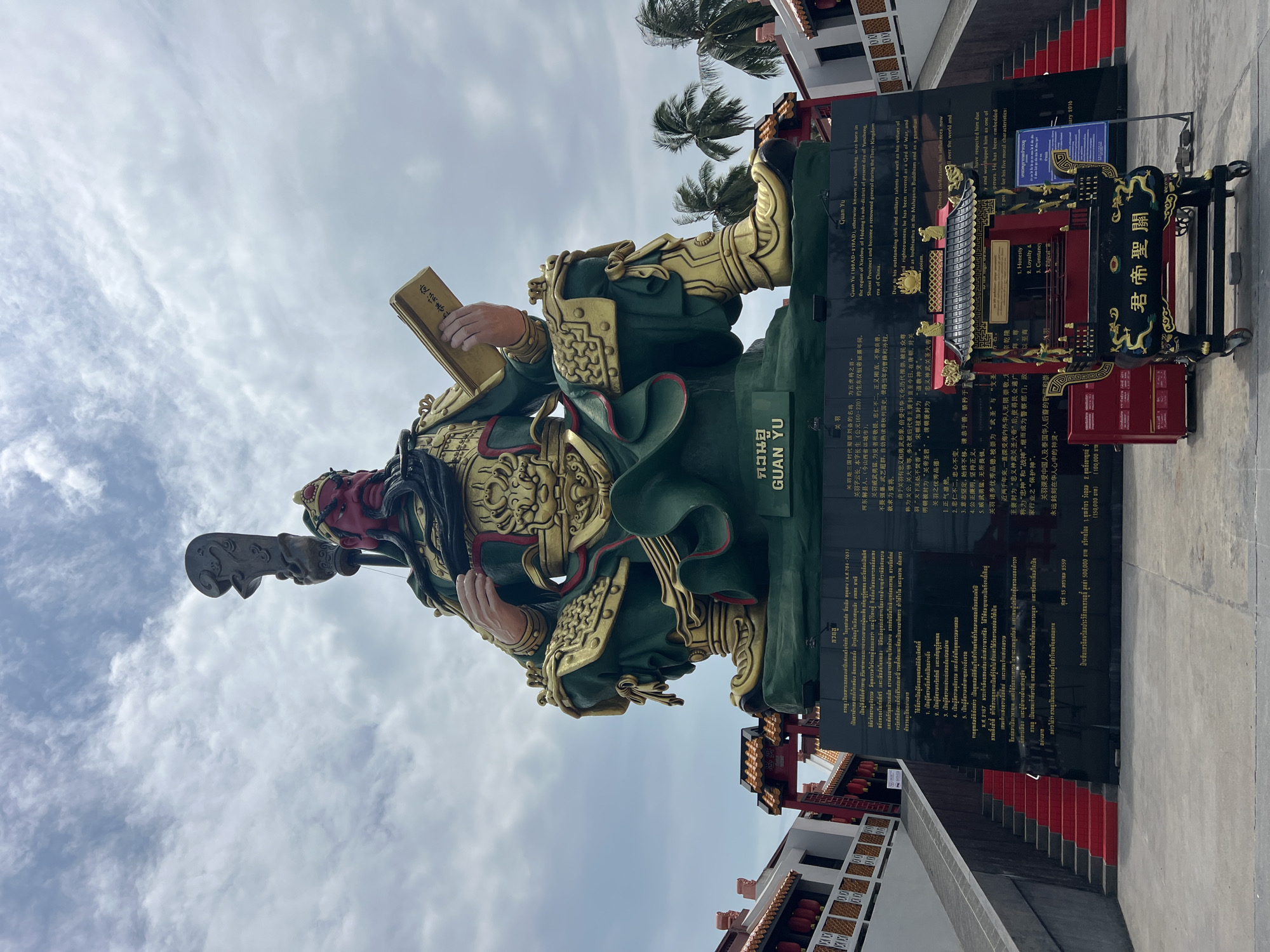 The image captures a statue of Guan Yu in Baan Hua Thanon, Thailand, set against a partly cloudy sky. The statue is a mix of green and gold, with Guan Yu holding a book in one hand and a weapon in the other. His face is painted red.
Behind the statue is a dark slab inscribed with text in multiple languages, including Thai and Chinese. To the right of the slab is a smaller ornate, red and black structure.
The statue is placed on a platform with a few steps leading up to it. A building with red accents is visible in the background. Palm trees are also visible. The statue is the main focal point and appears to be newly constructed and well-maintained.