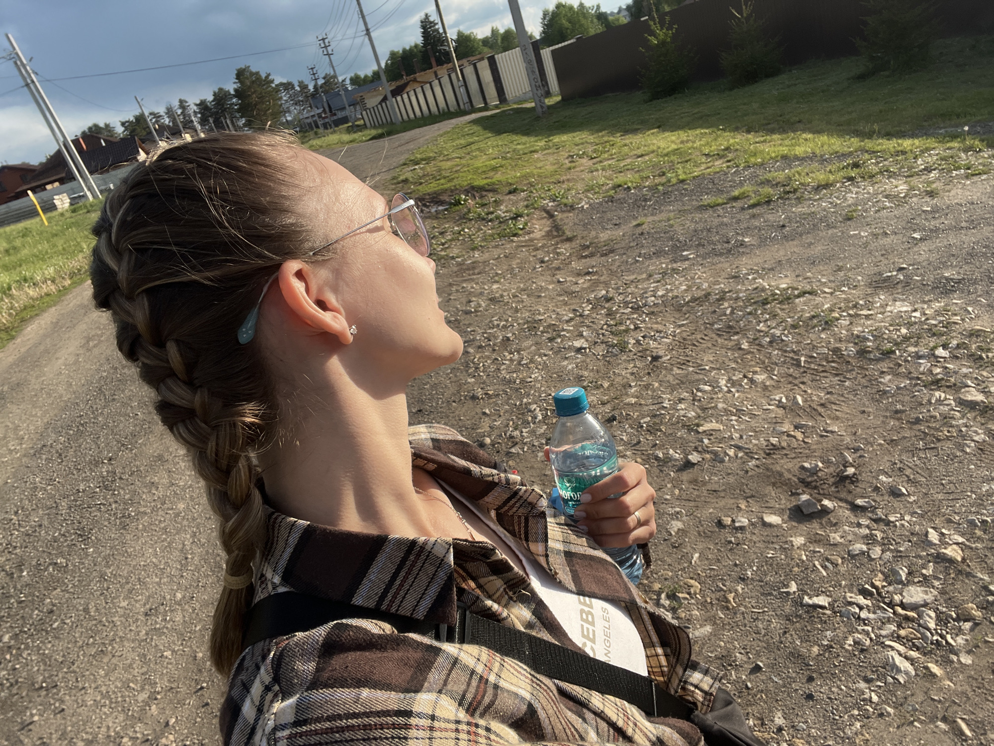 A woman with blonde hair braided into a single plait over her left shoulder is standing on a gravel road. She wears sunglasses, a brown and white plaid shirt over a white T-shirt with the words "CEBE ANGELES", and a black strap is visible across her torso. In her right hand, she holds a clear water bottle with a blue cap and a label in a Cyrillic script. The woman is looking up and to the right. The road is lined with green grass and trees. In the background are houses and a metal fence, as well as telephone poles. The lighting indicates it is daytime with bright sky conditions. The location is Chaikovsky, Russia.