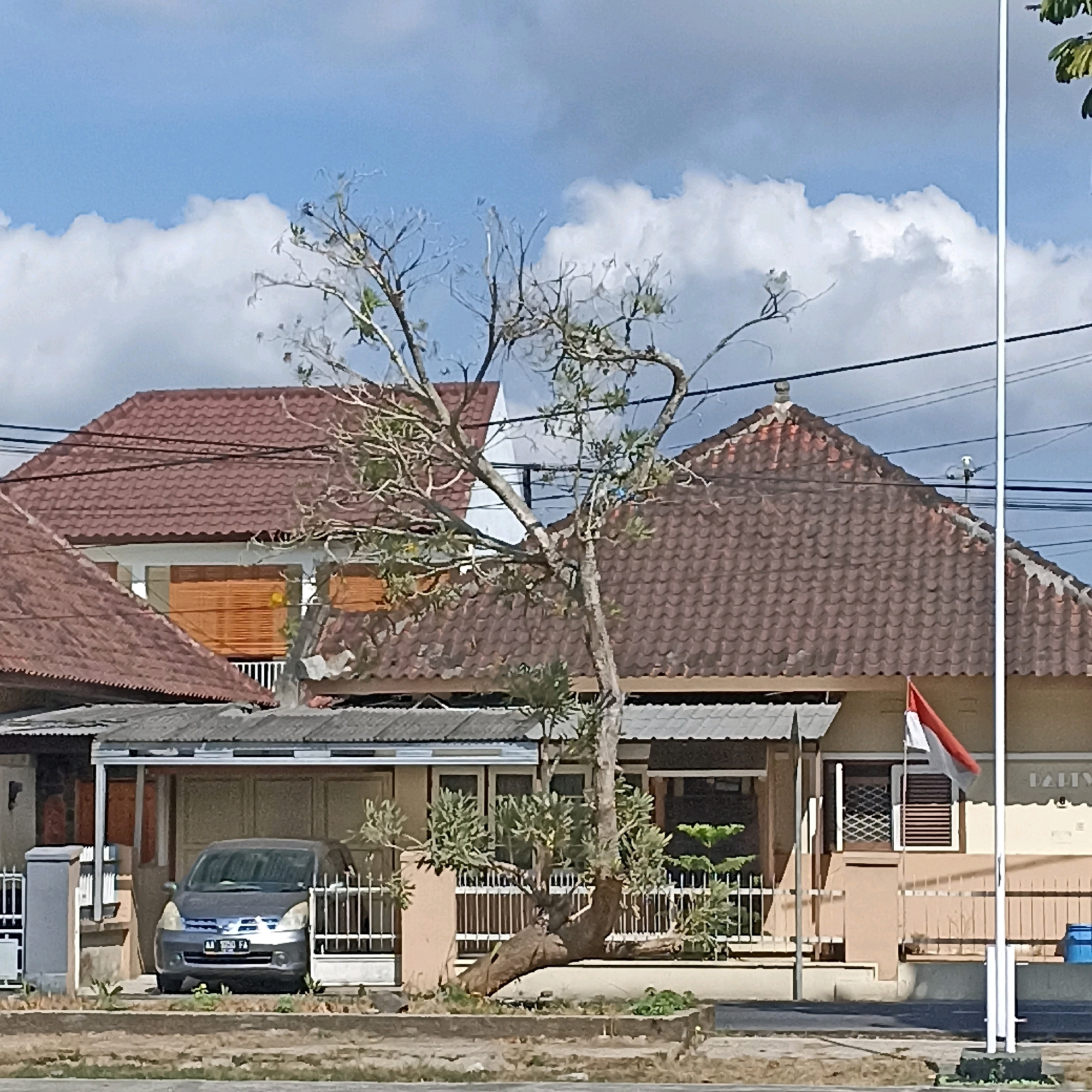 In the city of Magelang, Indonesia, a two-story house with a red tiled roof stands under a cloudy blue sky. A bare tree with a thick, twisted trunk occupies the foreground, its branches reaching towards the sky. To the left of the house, a grey car is parked under a covered carport. A small Indonesian flag, red and white, flutters on a flagpole in front of the house. The scene appears to be a residential area, with utility wires running overhead. The image is taken during daylight hours, with the sun casting shadows on the ground.