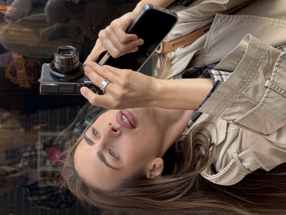A young adult female is depicted in a medium close-up, angled slightly upwards, outdoors in Venice, Italy. She has long, light brown hair, light-colored eyes, and light pink lipstick, with her mouth slightly open. She wears a light tan trench coat over a blue and brown plaid shirt. On her left ring finger, she exhibits a silver-toned ring with a screw-head motif pattern. Her gaze is directed upwards and to the right, consistent with aiming a photographic device. She holds a black Canon PowerShot G7 X Mark II compact digital camera in her left hand, with her thumb positioned near the shutter button. Her right hand supports a smartphone in a clear case, positioned beneath the camera. A brown leather strap, possibly from a bag or phone accessory, is visible over her right shoulder. The background is a blurred, dark, reflective surface, likely a shop window, displaying indistinct reflections of other individuals and fragments of a sign with orange lettering, indicative of a commercial street setting. The scene is illuminated by natural sunlight.