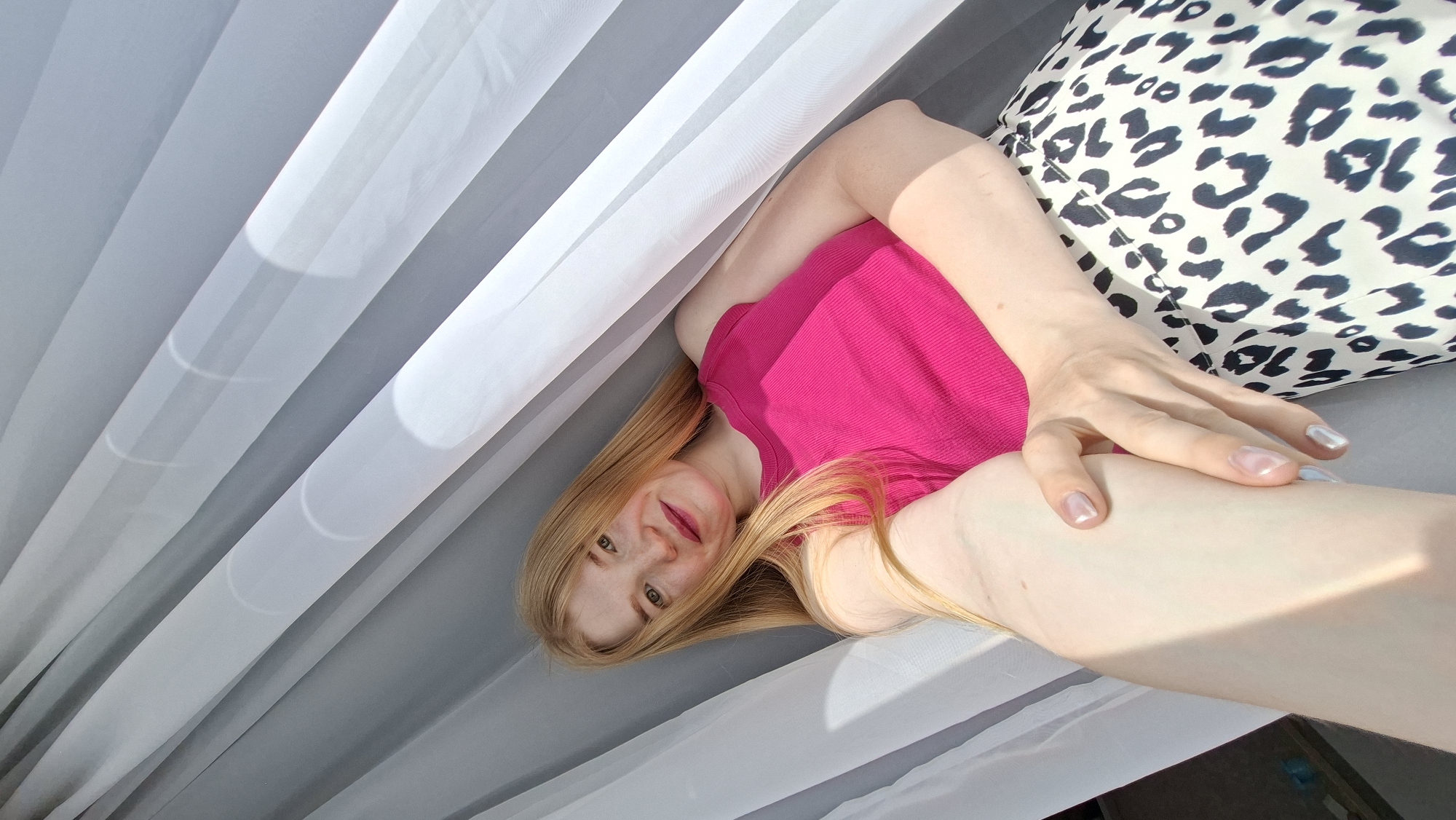 A woman with long blonde hair and fair skin is lying down indoors. She is wearing a bright pink top and what appears to be a leopard print garment beneath her. Her left arm is bent and resting on her leg. She has light-colored fingernails. She is positioned beneath a white and grey striped fabric, which may be a curtain or canopy. The location of the photo is Chaikovsky, Russia.
