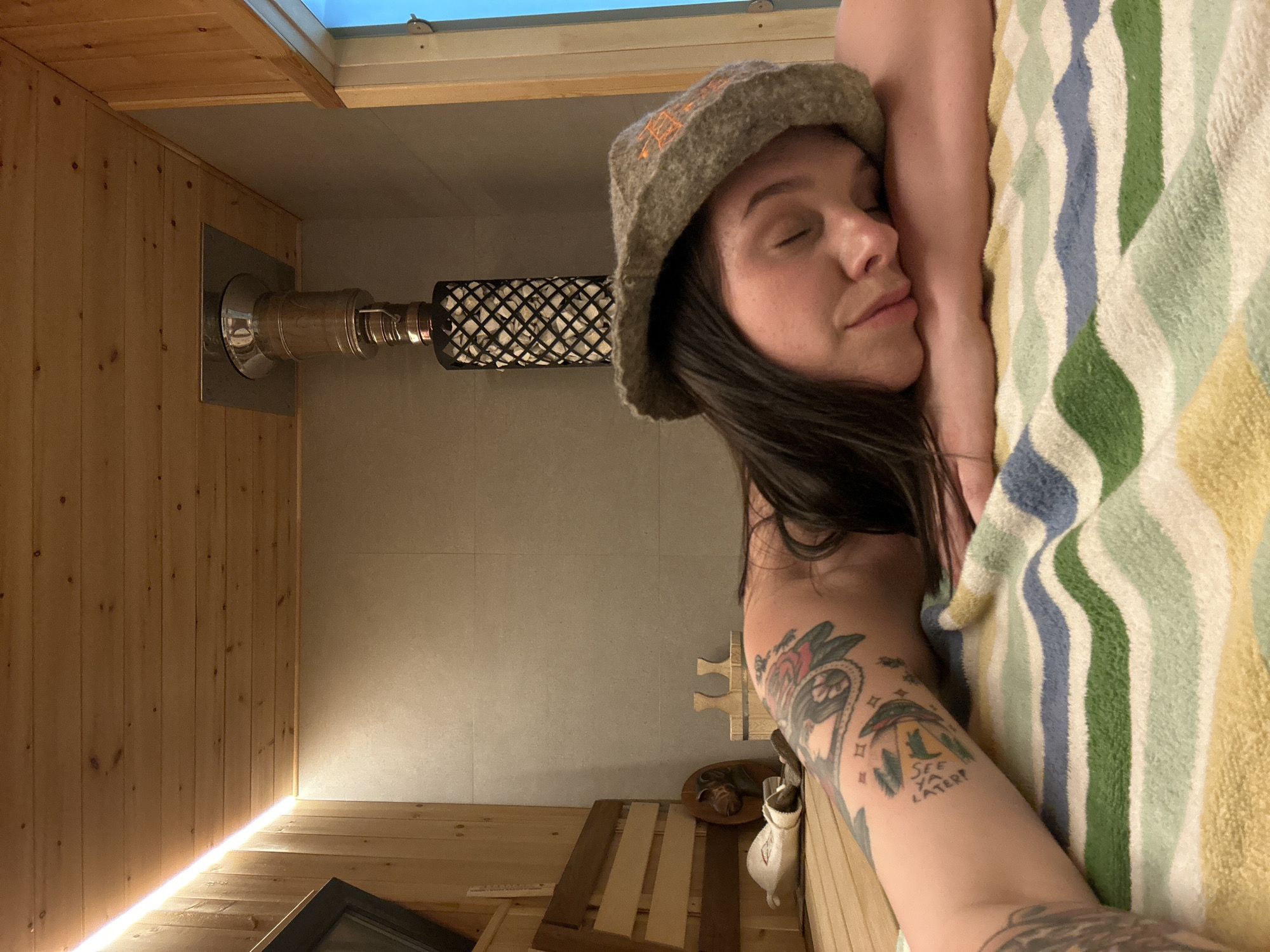 A woman wearing a sauna hat is lying with her eyes closed, partially covered by a striped towel. She has tattoos on her arm. She is in a sauna with wood paneling and a metal heater. The city of Прикамский, Russia is not discernable from the image.