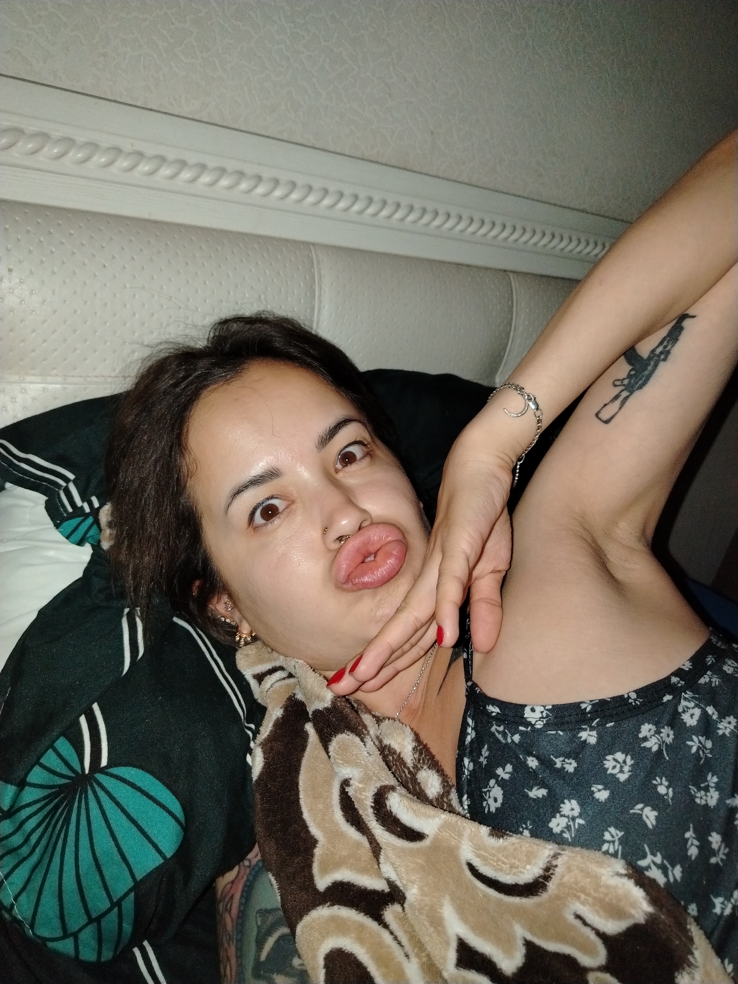 A woman with dark hair and brown eyes lies in bed, making a kissing face. She is wearing a black dress with a floral pattern and a patterned blanket is draped over her chest. Her right arm is raised, showing a tattoo of a rifle on her upper arm and a bracelet on her wrist. Her left hand is near her face, with red nail polish visible on her fingernails. The bed has a white upholstered headboard. The scene is indoors, likely a bedroom.